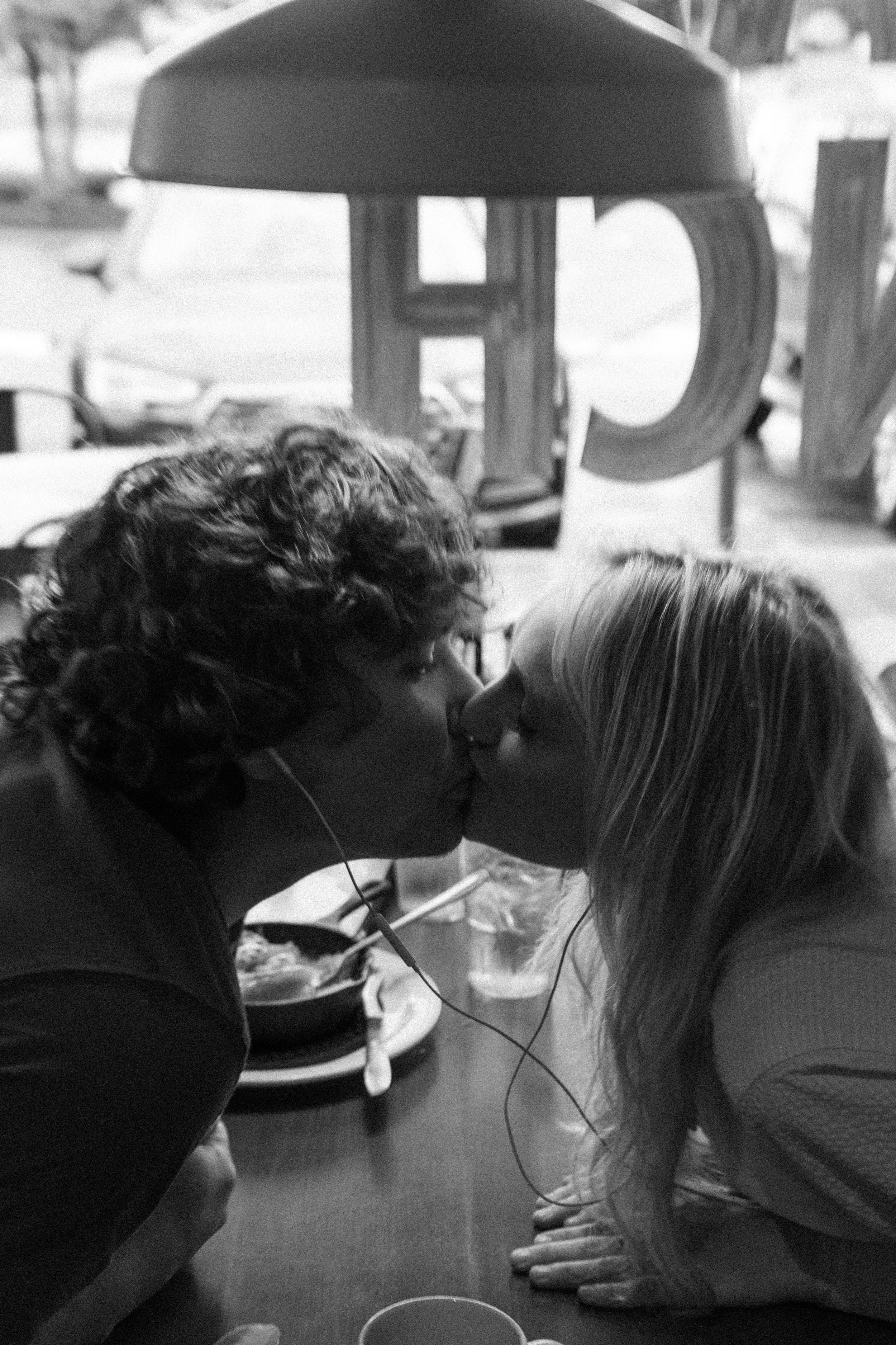 A black and white photo of a man and woman kissing across a table in a casual restaurant. The man has curly hair and is wearing earphones, while the woman has long straight hair. In the background, large letters spell out part of the word 'LOVE'.