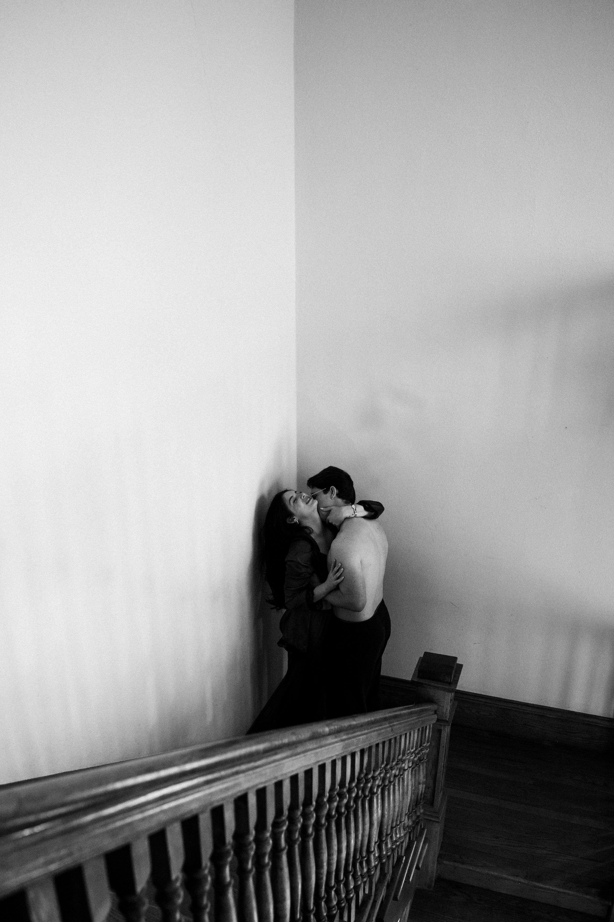 A black and white photo of a shirtless man embracing and kissing a woman against a wall on a staircase.