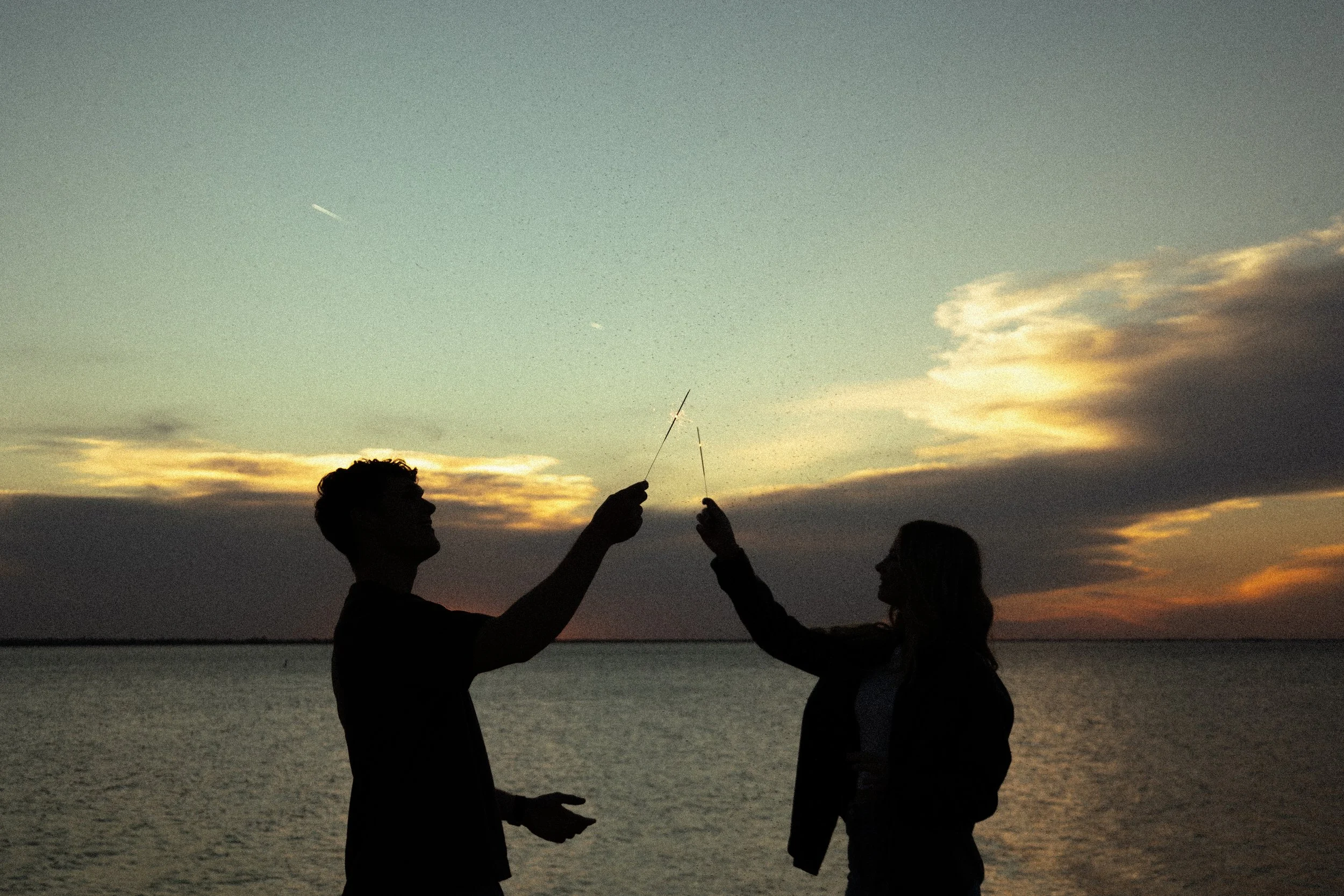 Silhouettes of a young man and woman by the water during sunset, holding sparklers in the air.
