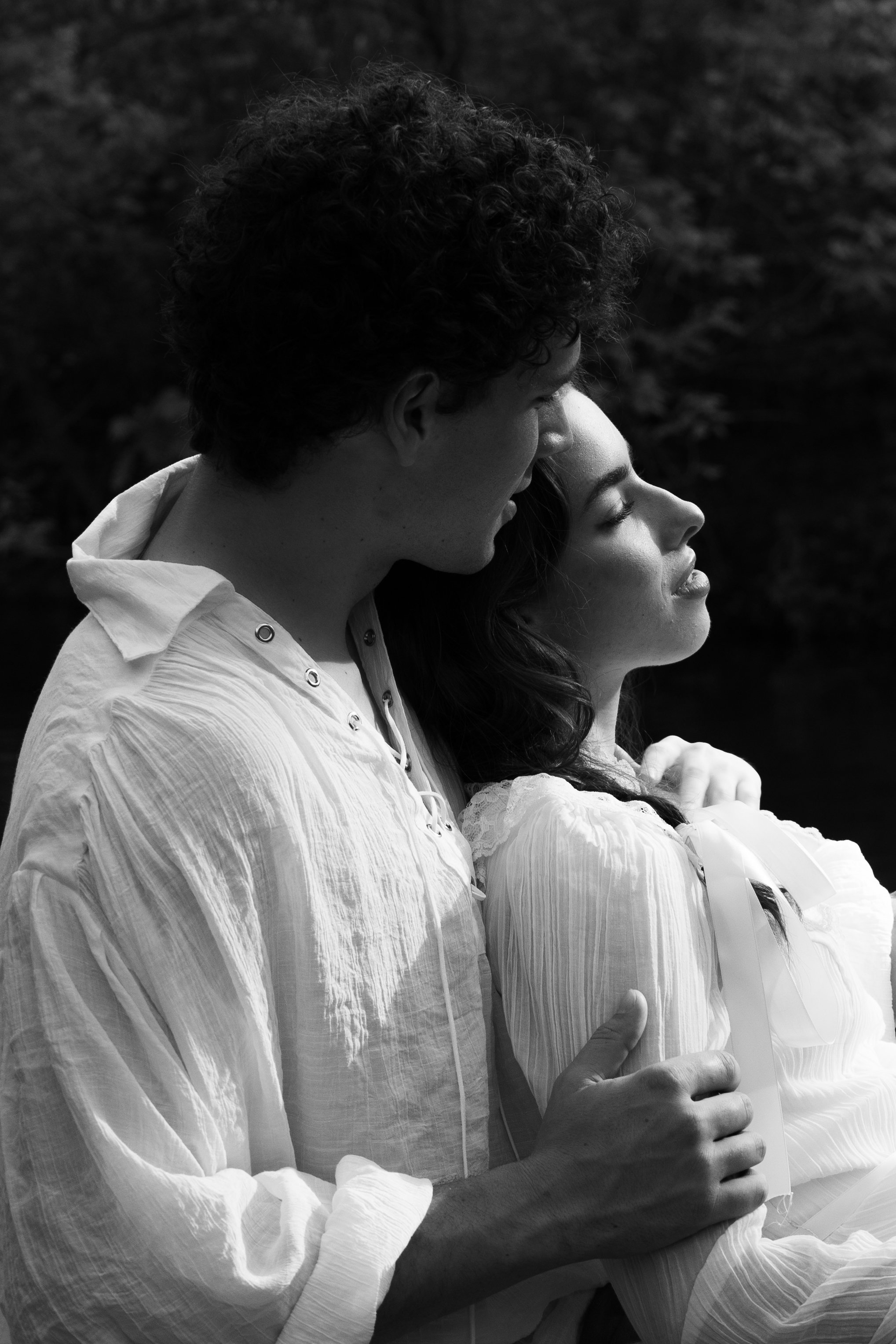 A black and white photo of a man and woman embracing outdoors, with the man's face close to the woman's forehead. The man has curly hair and is wearing a light-colored, slightly crinkled shirt. The woman has long dark hair, her eyes closed, and is we