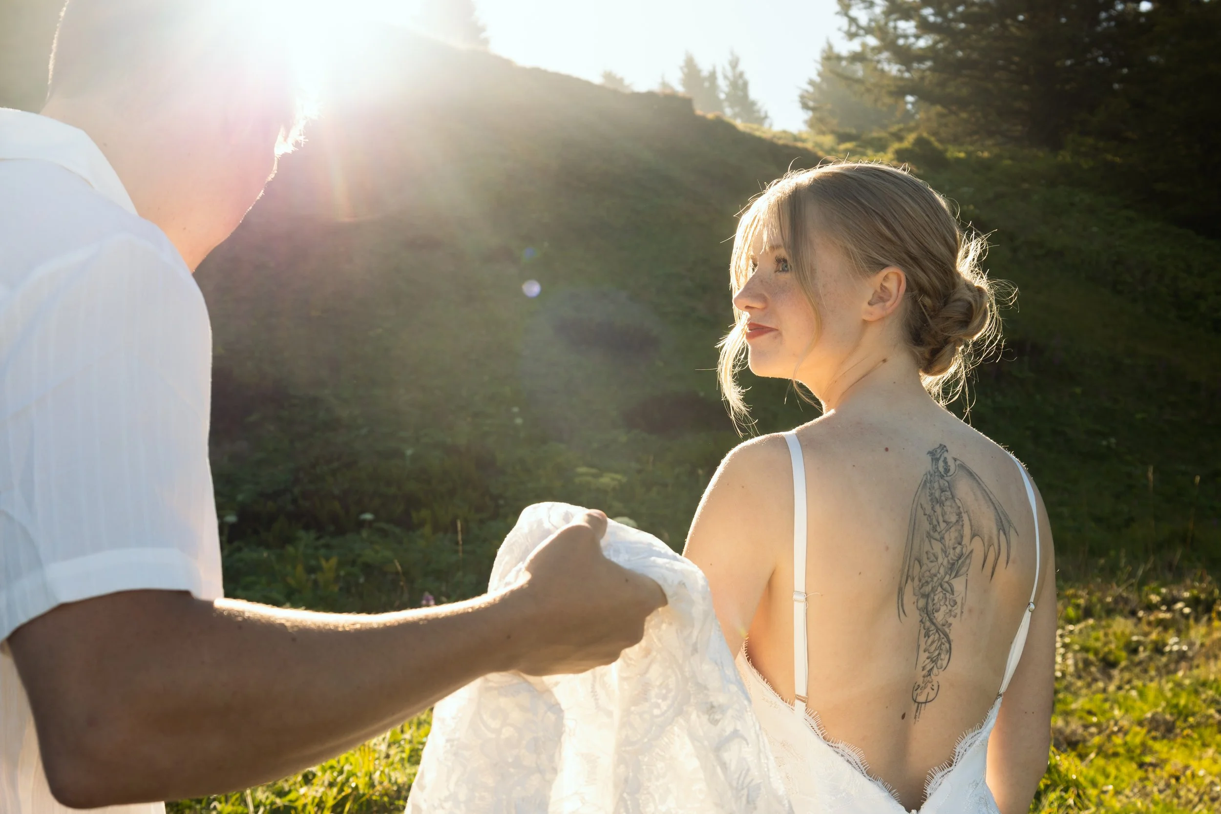 A woman with a tattoo on her back and light-colored hair styled in an updo, sitting outdoors during sunset, while a person in a white shirt adjusts her dress.