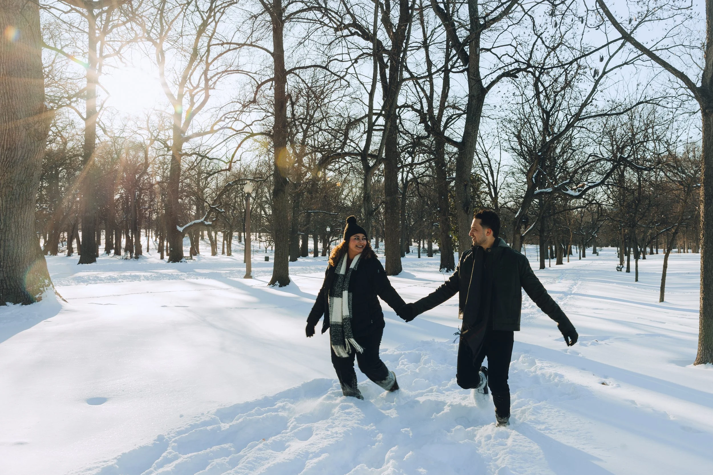 A couple holding hands and walking in a snow-covered park with leafless trees and sunlight shining through