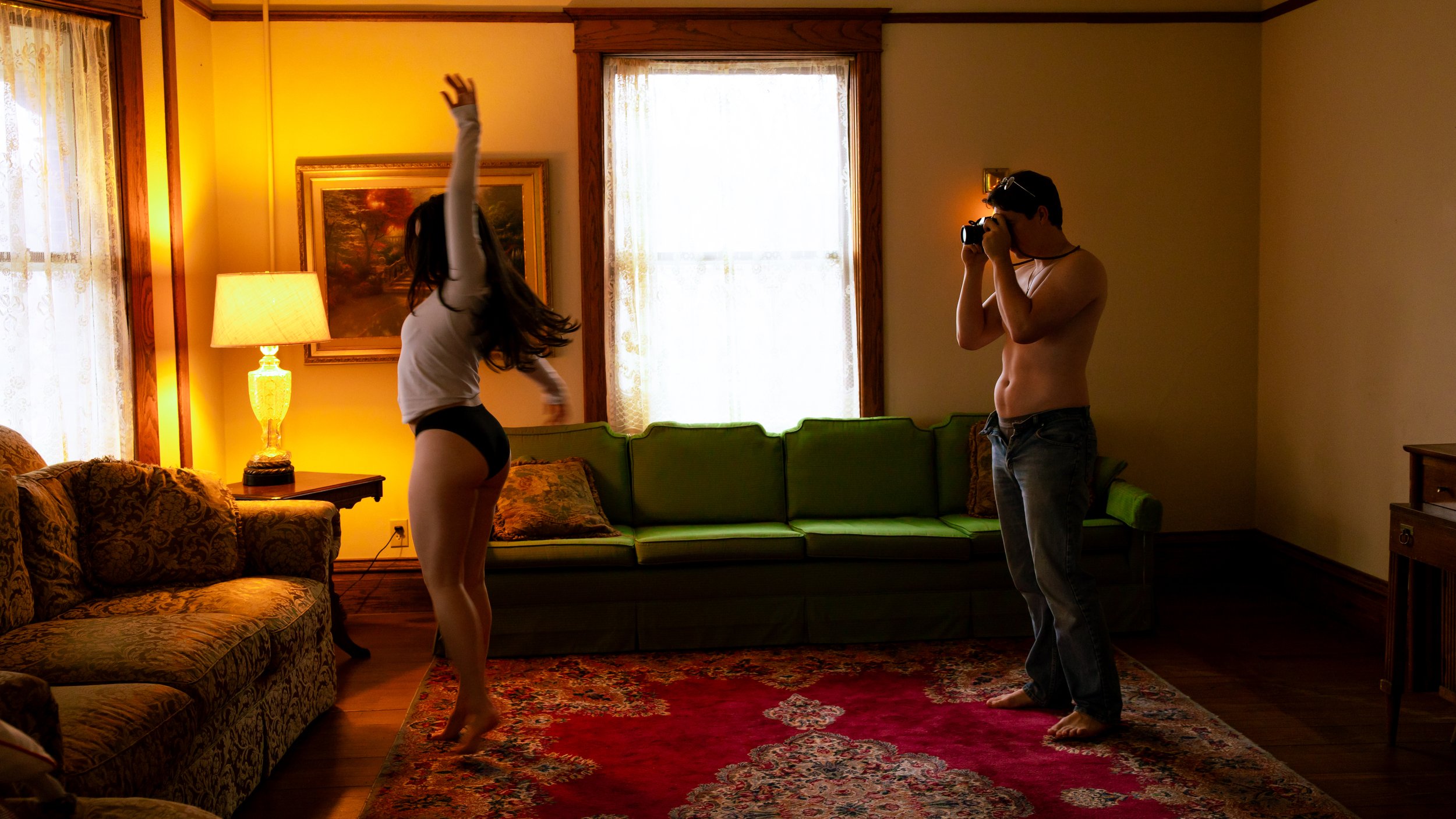 A woman dancing in a living room while a man photographs her. The room has vintage furniture, warm lighting, and patterned rugs.
