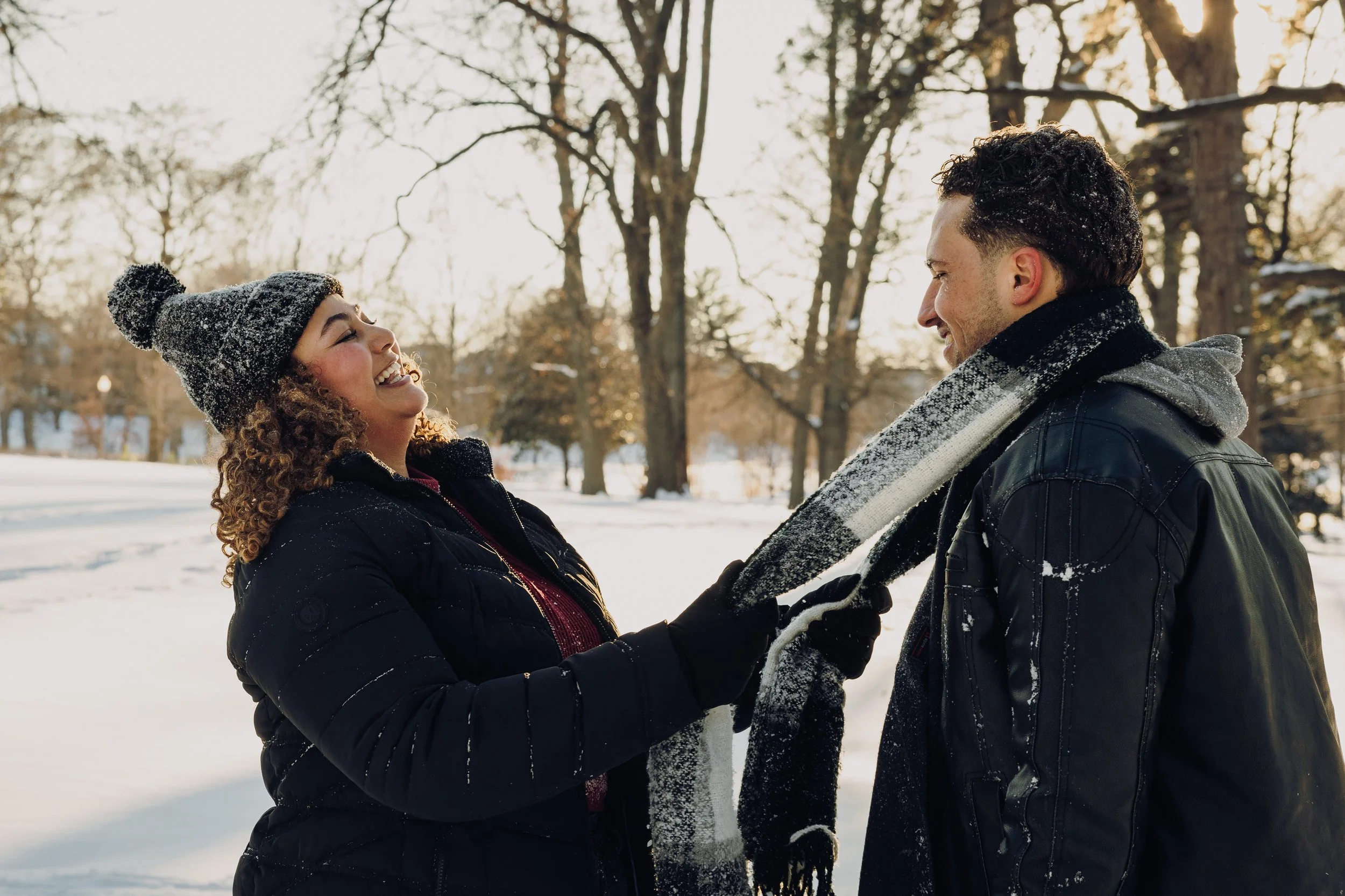 A woman and man laughing and smiling outdoors in a snowy park, with snow-covered trees in the background. The woman is wearing a black jacket, a knitted hat, and gloves. The man is wearing a black jacket and a scarf.
