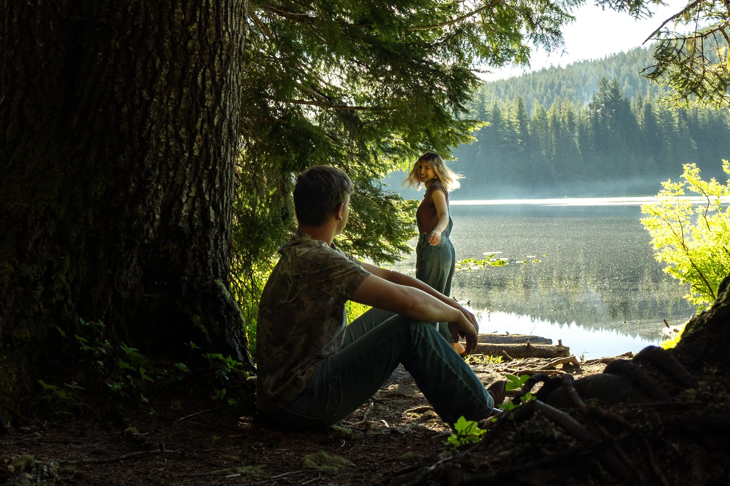 A man sitting under a large tree near a lake with a woman standing by the water's edge, smiling, surrounded by green trees and mountains in the background.