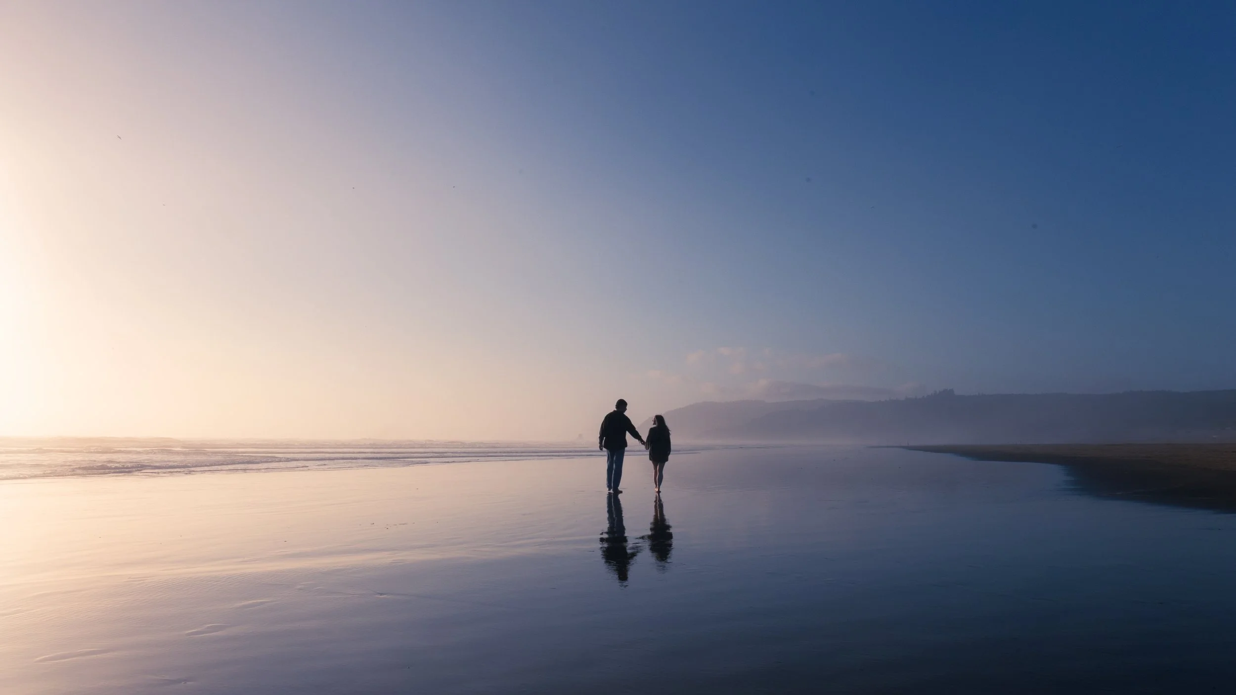 A couple holding hands walking along the shoreline on a beach during sunset or sunrise, with reflections on the wet sand and a distant mountain range under a clear sky.
