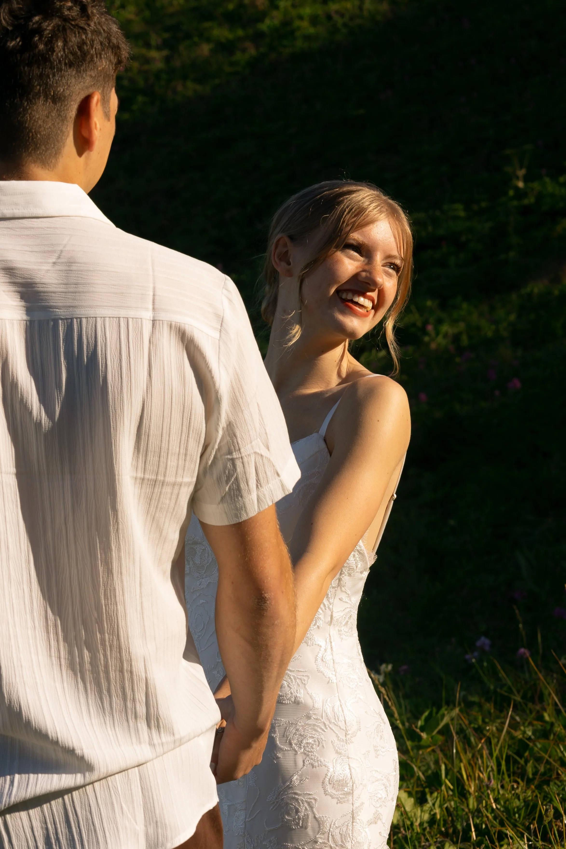 A young woman in a white dress smiling and looking at a man in a white shirt outside during golden hour.