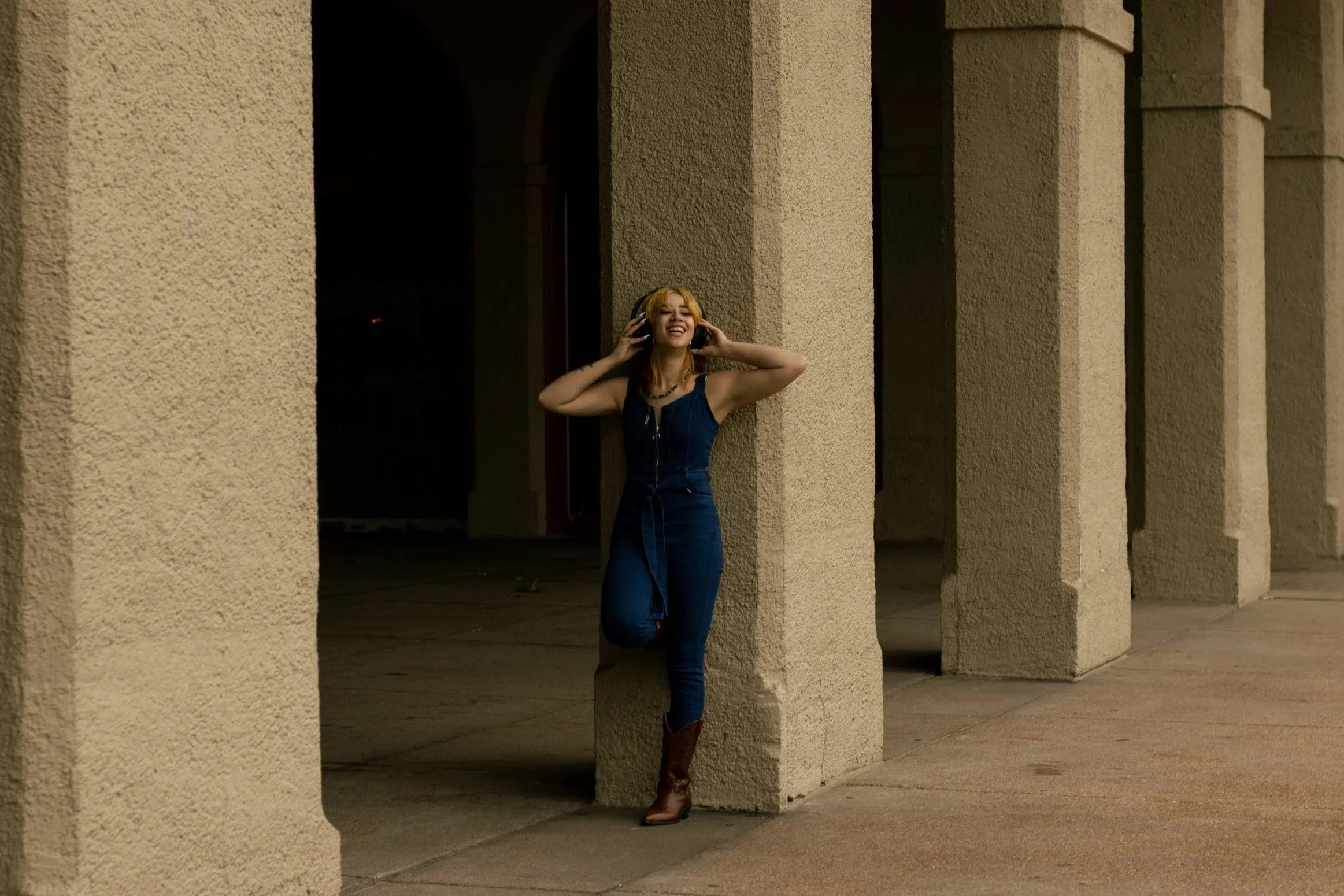A woman with blonde hair, wearing a denim jumpsuit and cowboy boots, is standing against a beige textured wall with arches, smiling and listening to music with headphones.
