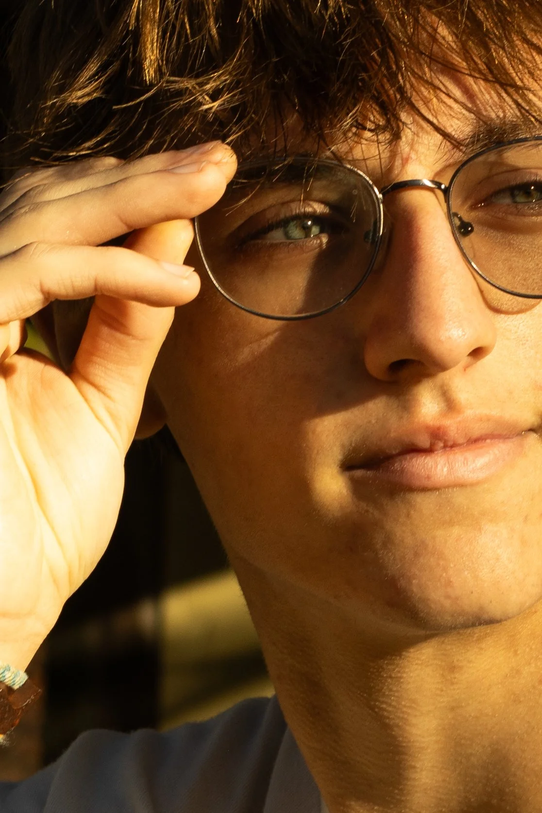 Close-up of a young person with glasses, holding the frame of their glasses with their right hand, looking at the camera in warm light.