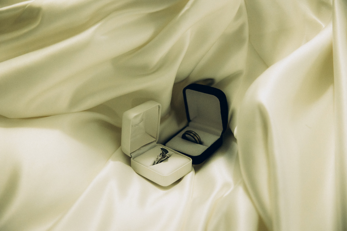 A wedding ring set inside a black jewelry box and an engagement ring inside a white box, both resting on a silky cream-colored fabric.