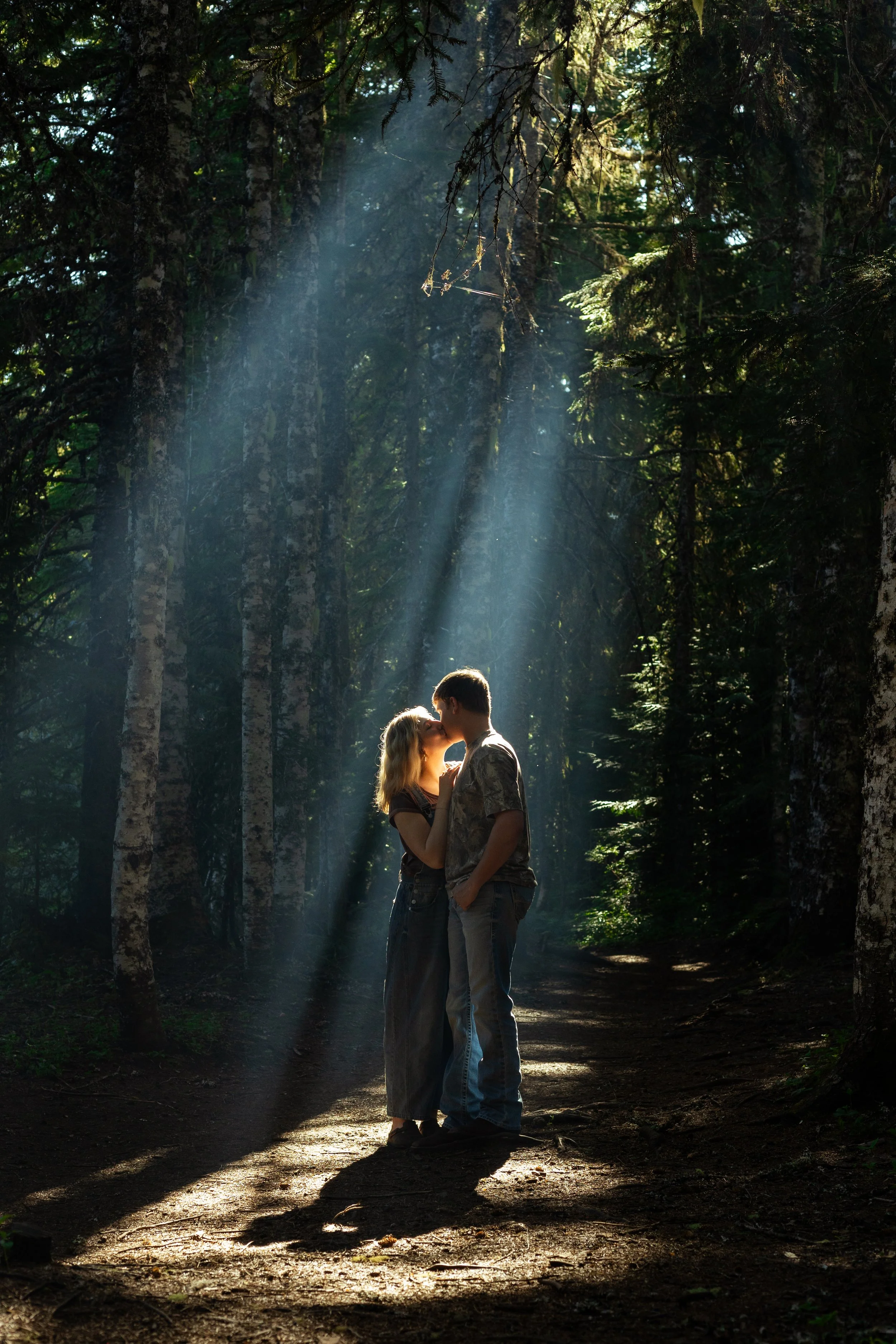 A couple sharing a kiss in a sunlit forest, with rays of light shining through the trees.