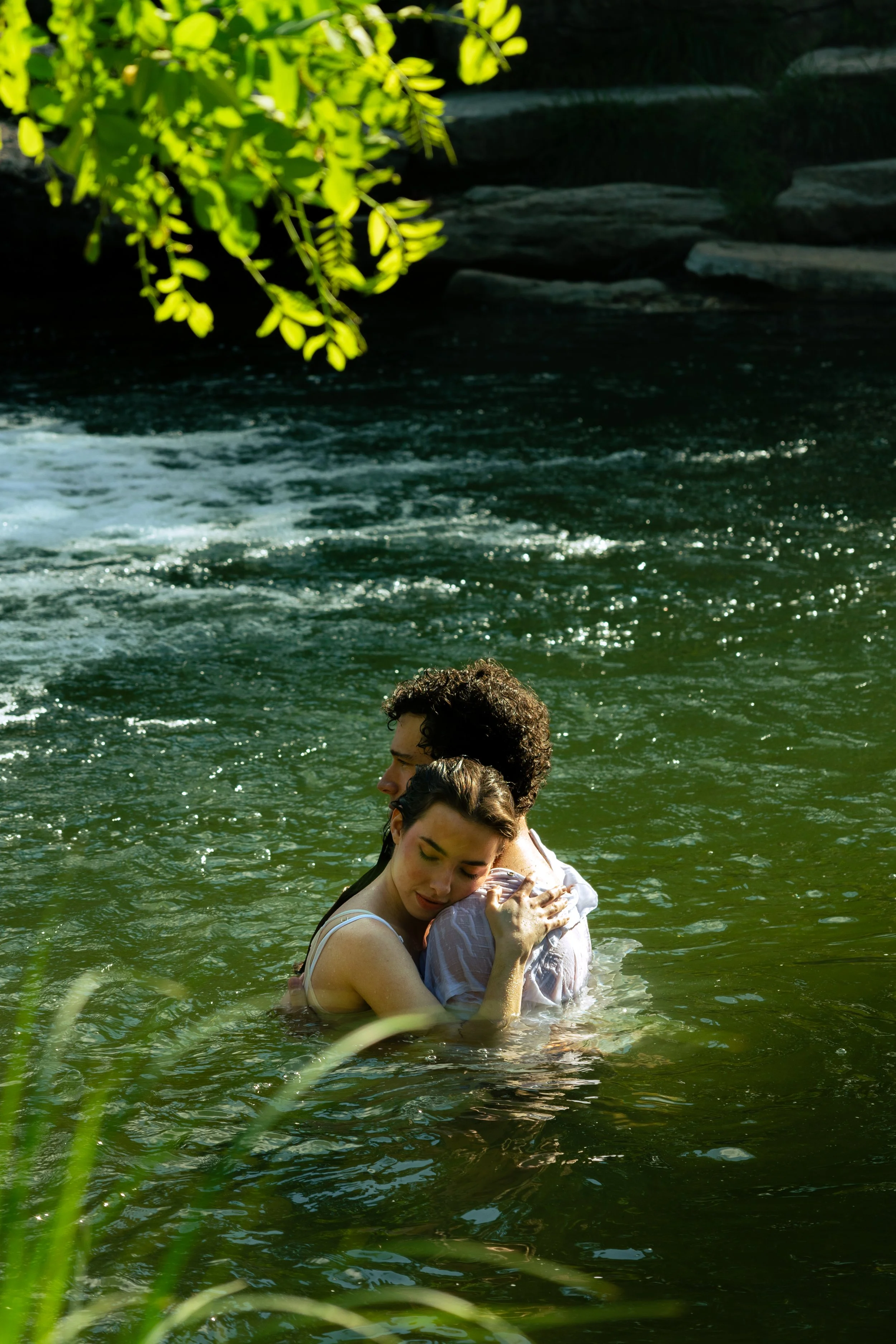 A young woman and man hugging in a river with greenery and rocks in the background.