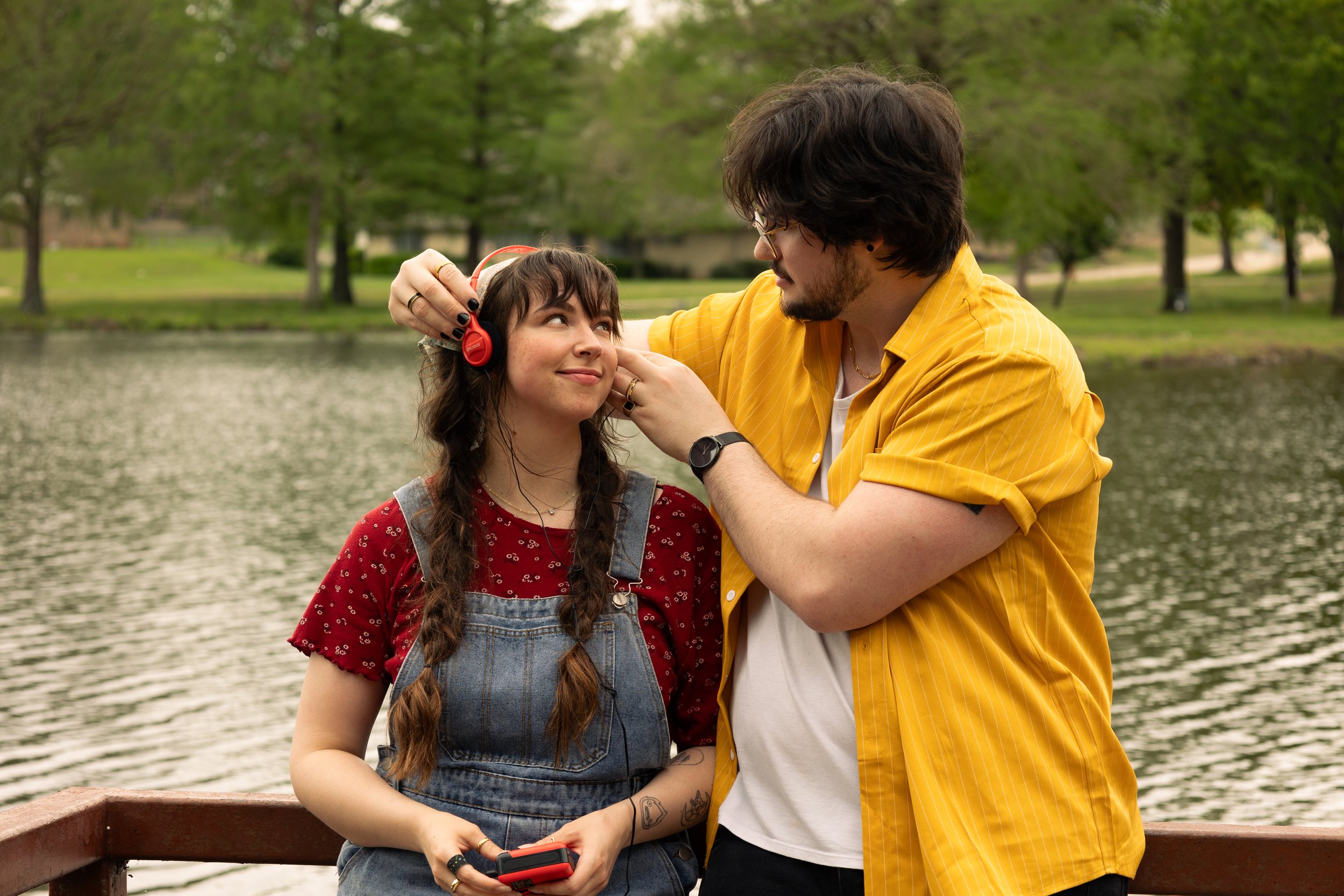 A young woman with glasses and long braided hair is listening to music with headphones, while a young man adjusts her headphones near a lake surrounded by trees.