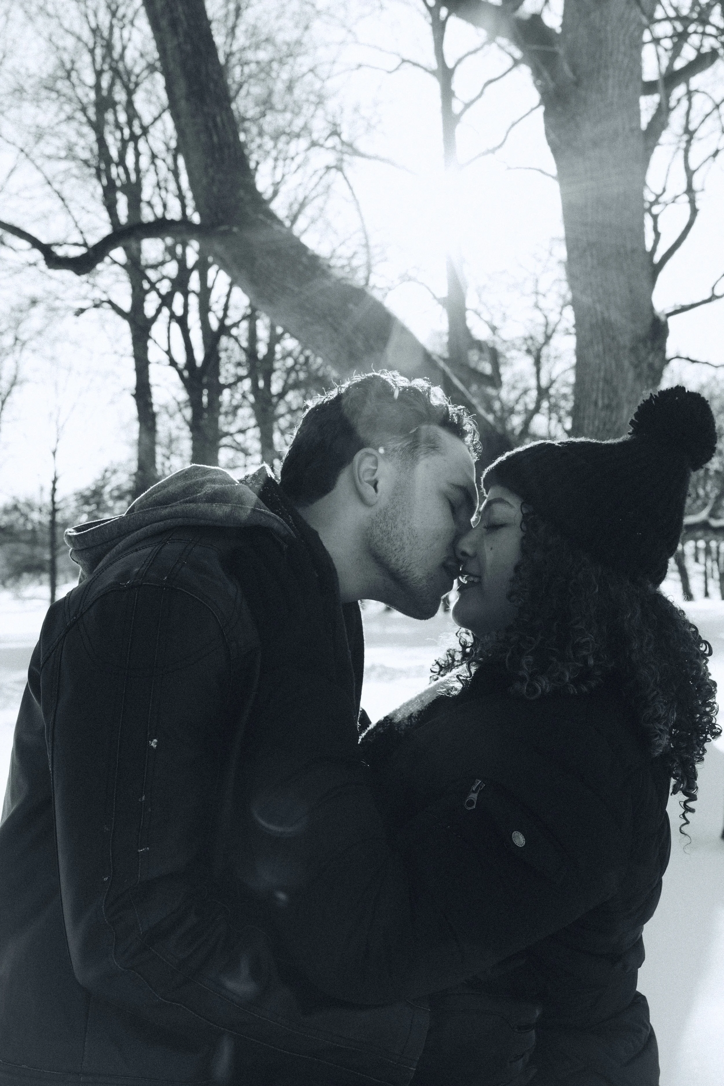 A black-and-white photo of a couple in a snowy park, sharing a kiss. The man has short hair and is wearing a jacket, and the woman has curly hair, a beanie, and a jacket. They are embracing each other, with trees and sunlight in the background.