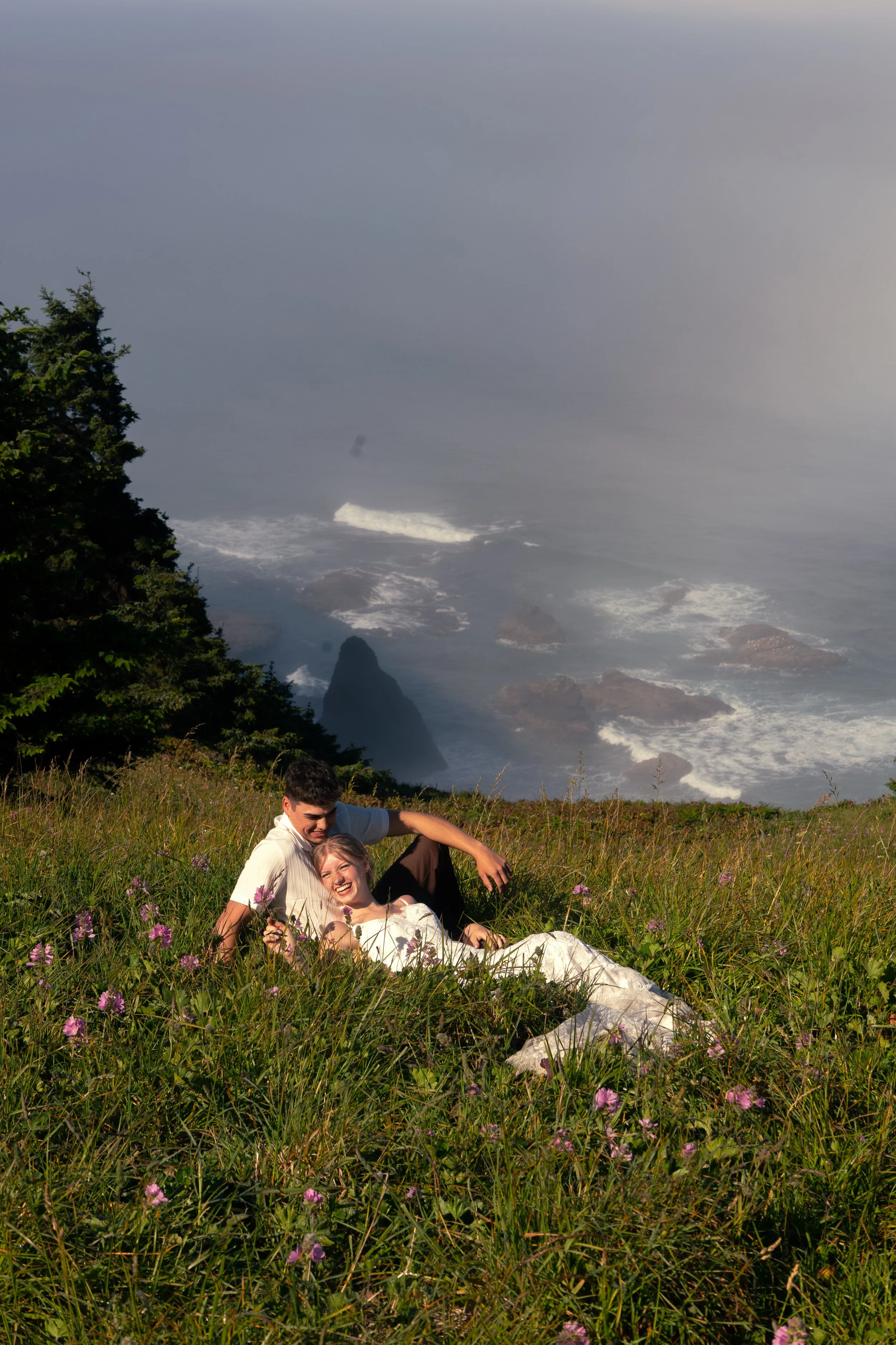 A couple lying on grass with purple flowers on a hill overlooking the ocean with rocks and waves.