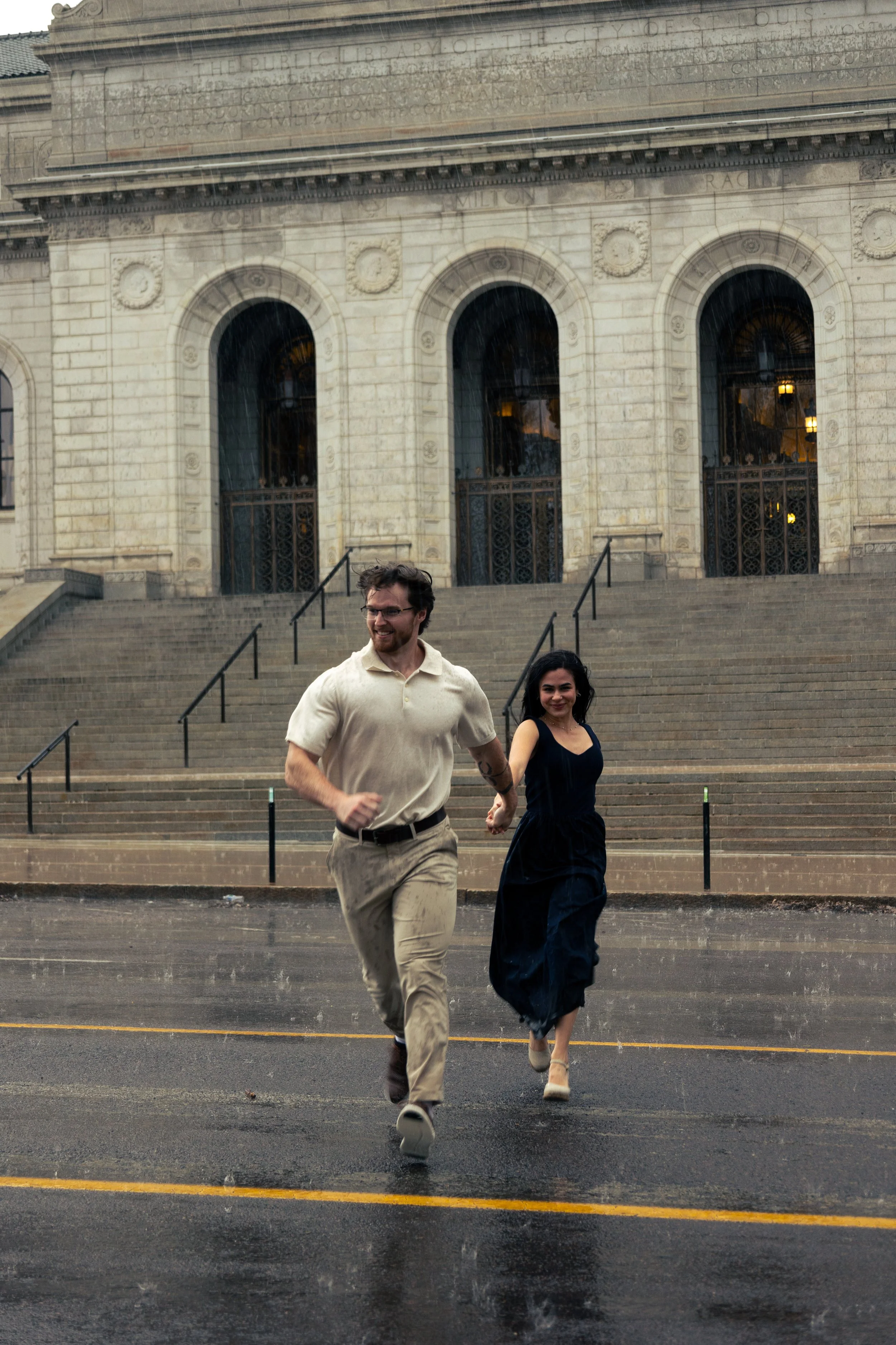 A young man and woman running hand in hand on a rainy street in front of a historic building with large arched windows and stone steps.