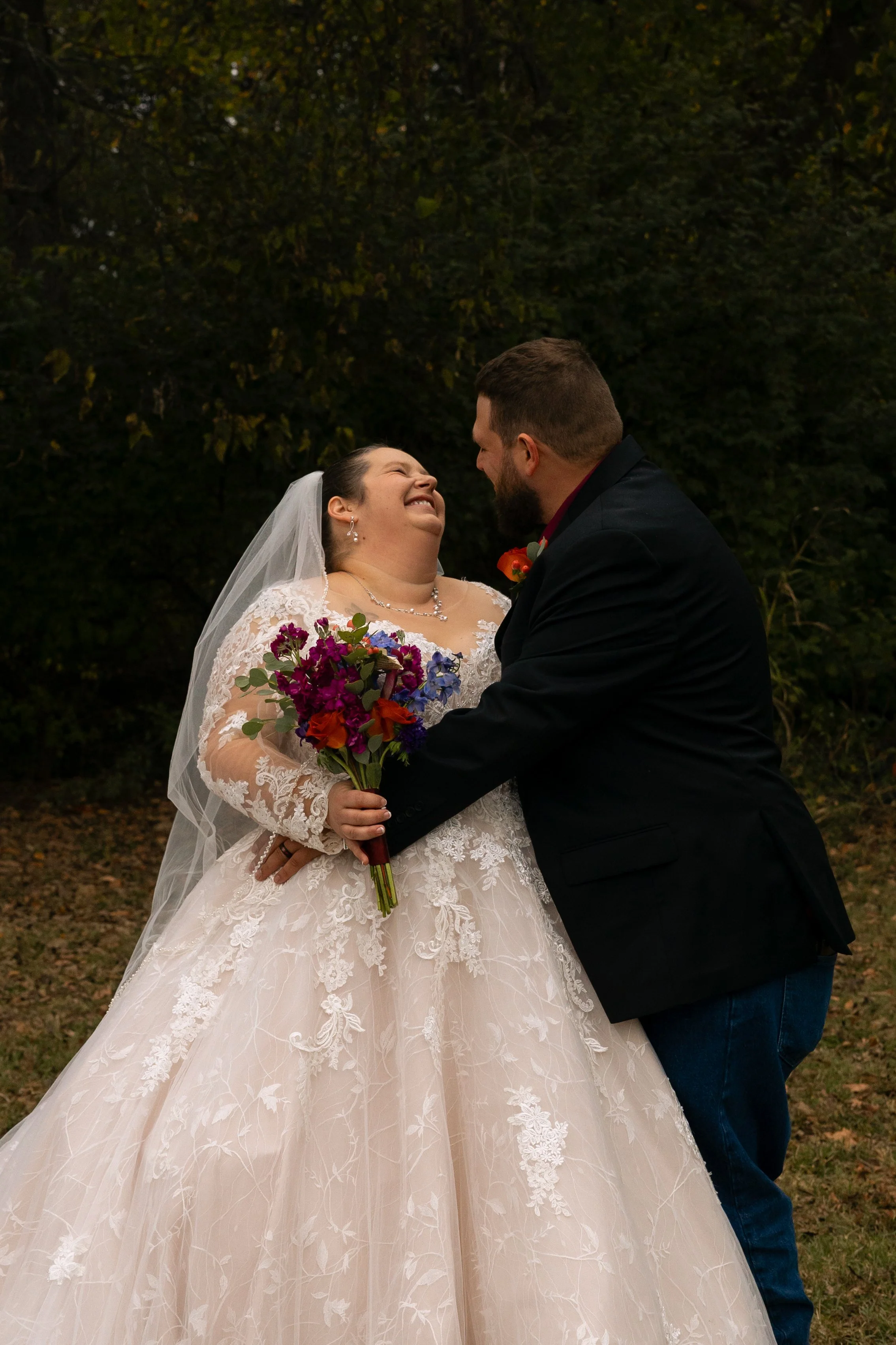 A bride and groom share a joyful moment outdoors during their wedding, with the bride holding a colorful bouquet and both smiling at each other.