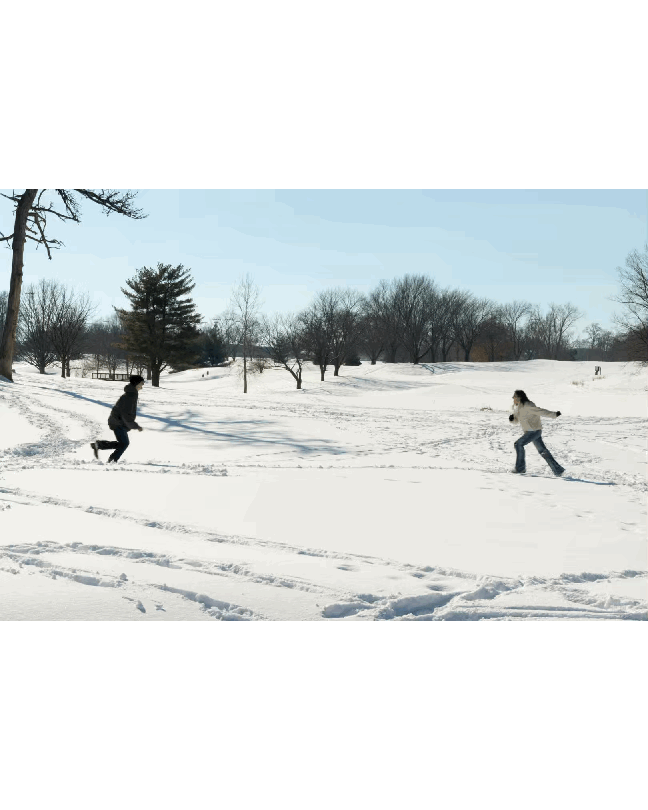 Two people are playing in a snowy park, one is running after the other. The park has snow-covered ground with scattered trees and a clear sky.