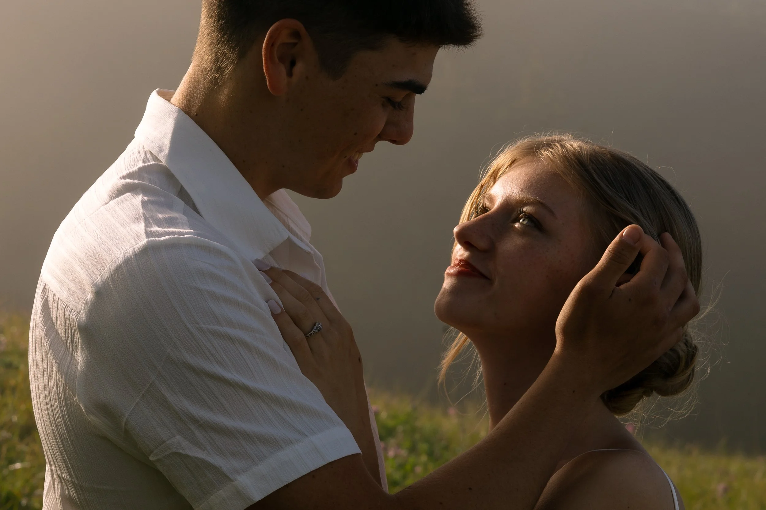 A young man and woman are gazing at each other lovingly outdoors during sunset, with the man holding the woman's face gently.