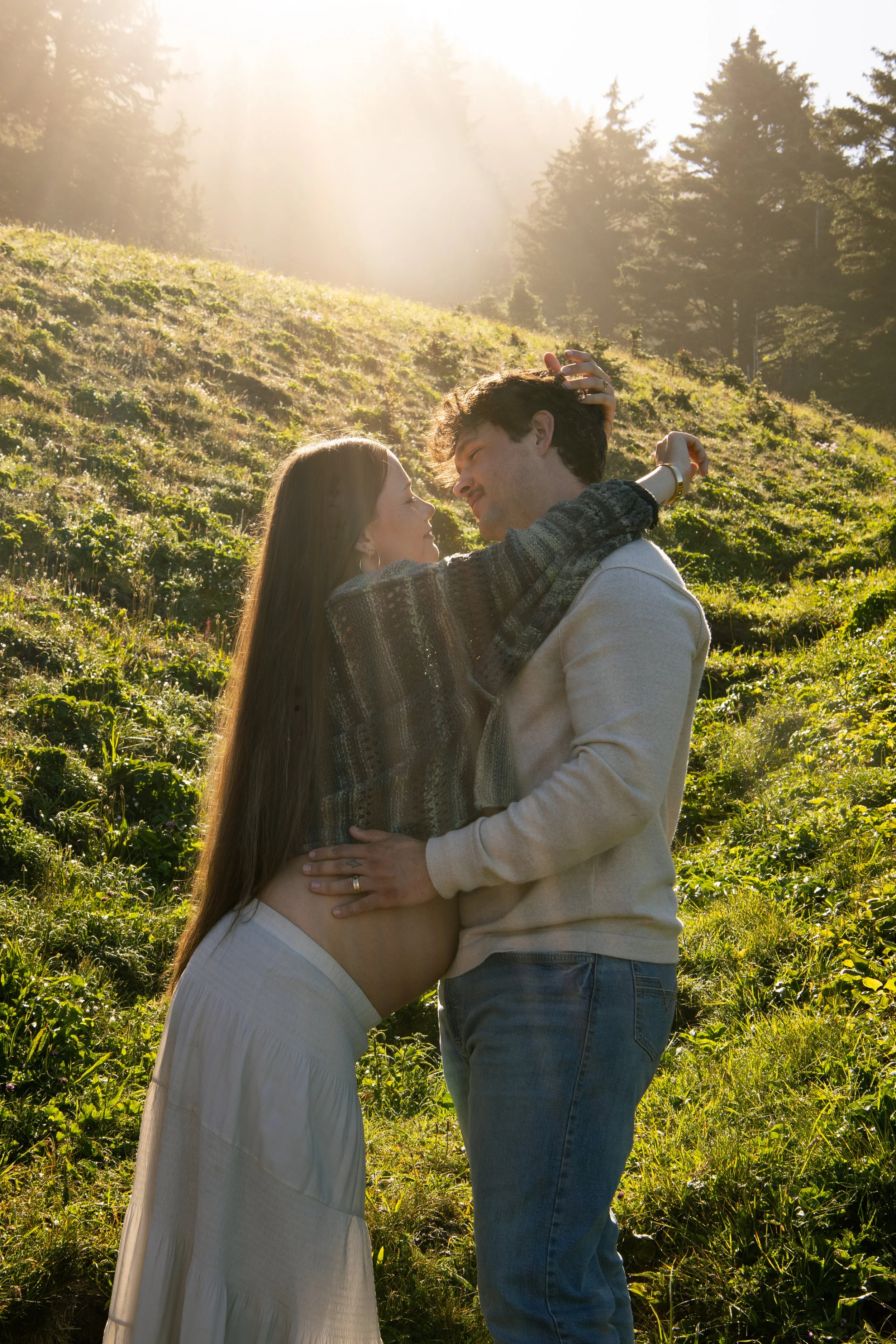 A couple embracing outdoors on a grassy hill with sunlight shining through trees in the background.
