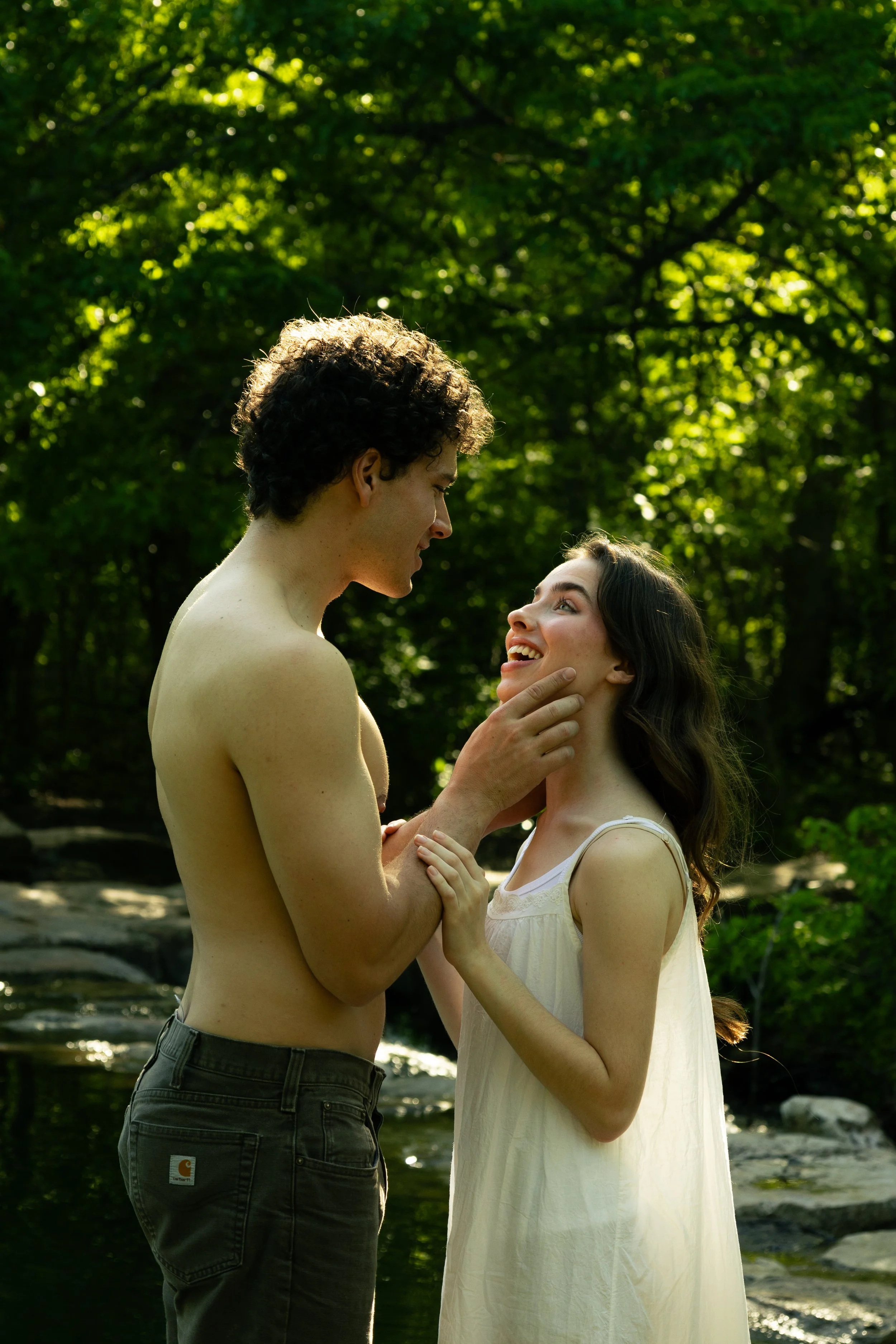 A young couple standing in a forest near a creek, smiling and gazing into each other's eyes, with the man touching the woman's chin.