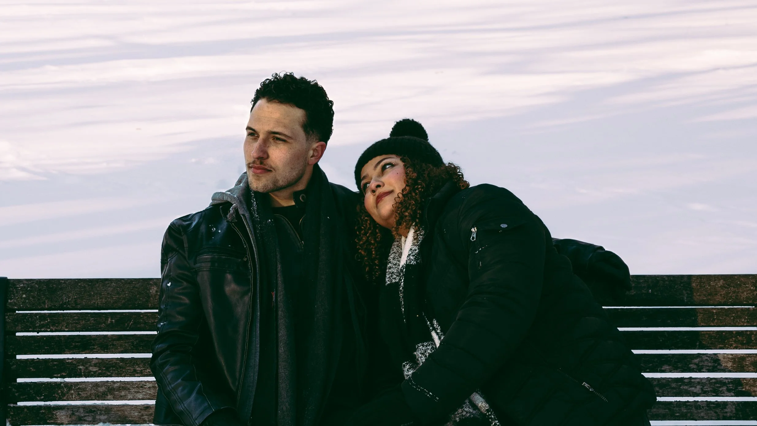 A man and a woman sitting on a park bench outdoors in winter, with snow on the ground and a cloudy sky in the background. The man has short dark curly hair, a light complexion, and is wearing a black leather jacket. The woman has curly hair, a darker