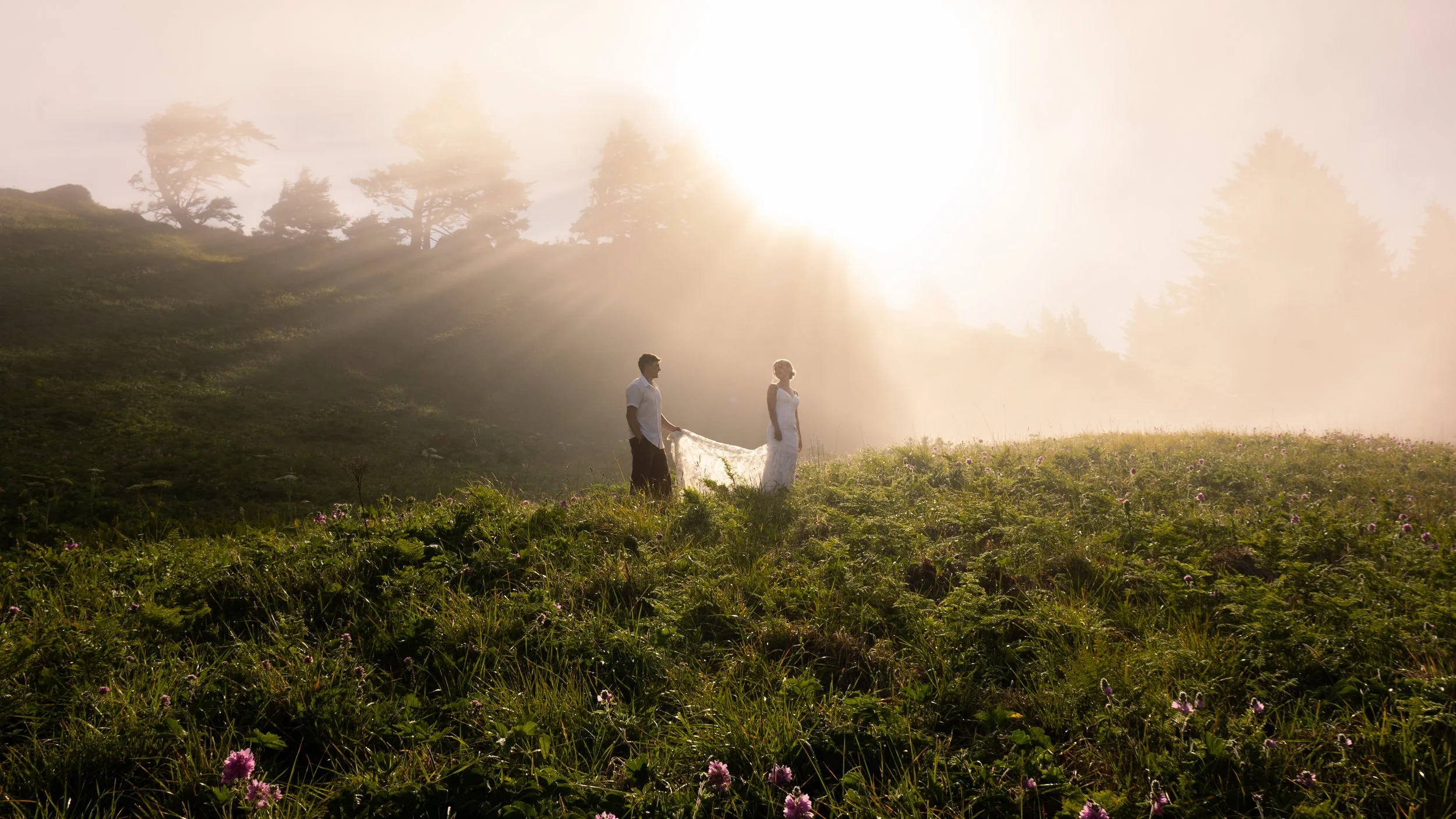 A couple in wedding attire in a lush green field during sunrise or sunset, with sunlight streaming through trees in the background.