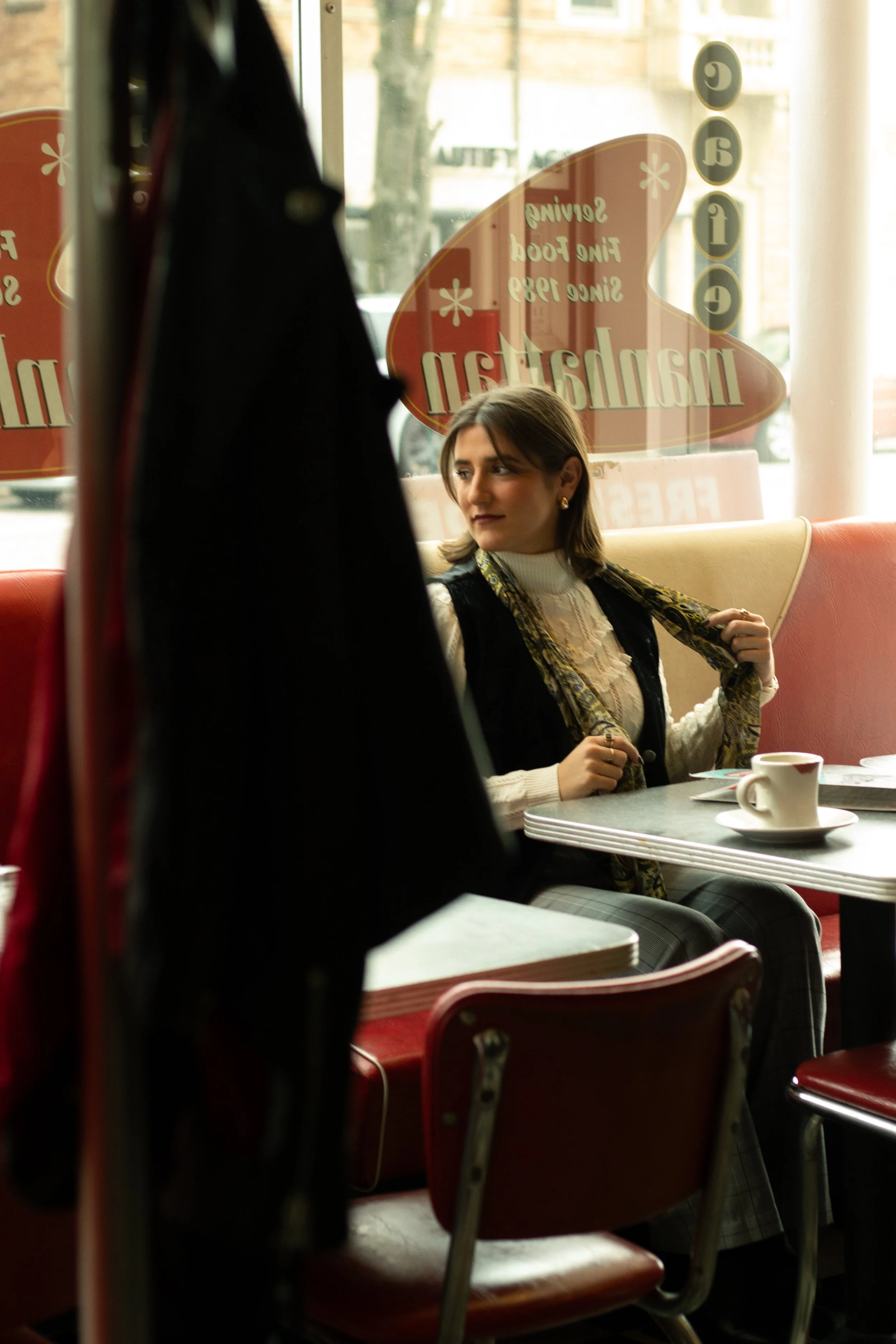 A woman with short brown hair, wearing a white turtleneck and a black vest, sitting in a vintage-style diner booth next to a table with a coffee cup and saucer, looking to her left, with a large window behind her reflecting outside scene and text on 