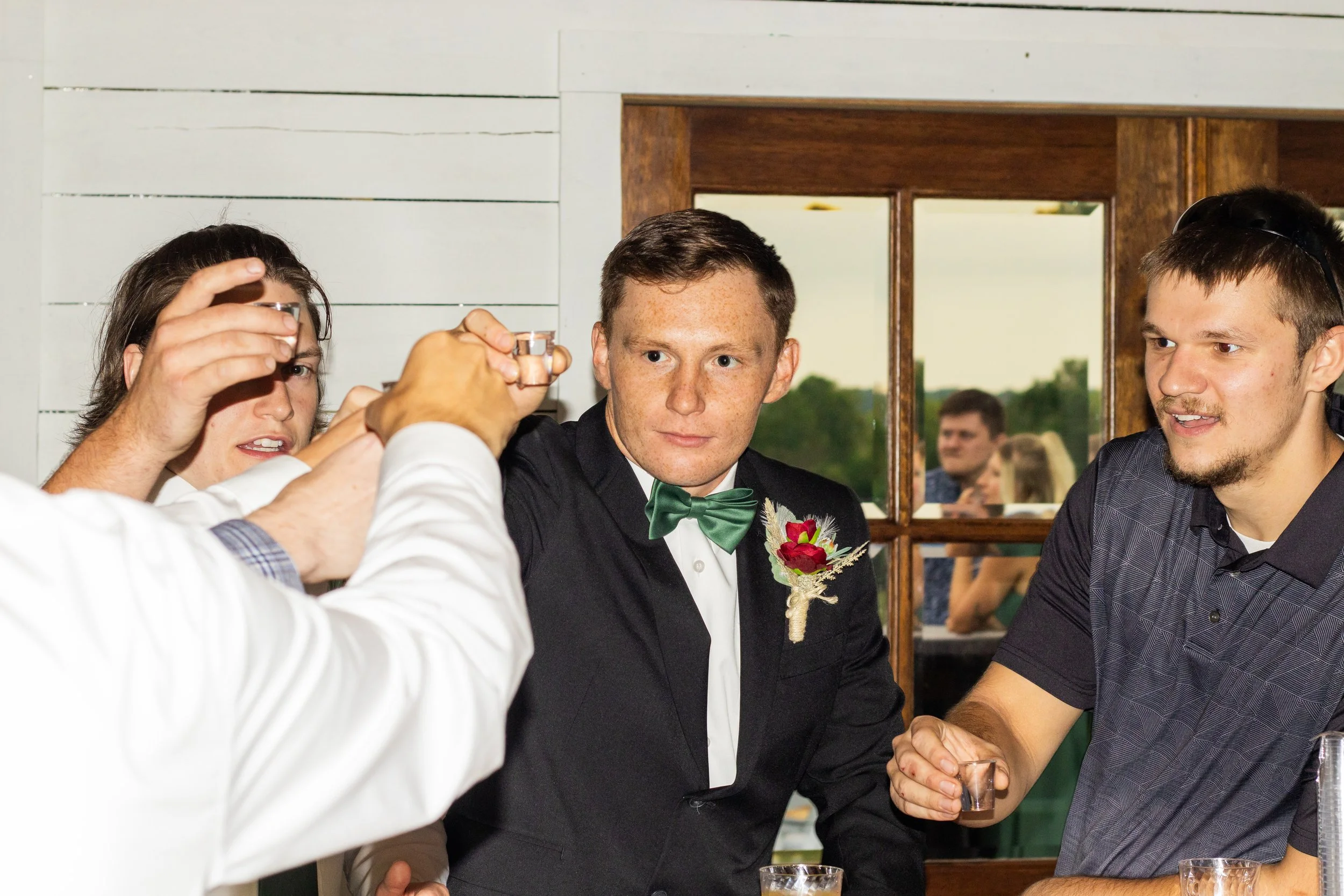 Group of young men at a celebration, one in a tuxedo with a green bow tie and boutonniere, friends toasting with shot glasses, standing near a wooden door with windows revealing outdoor greenery.