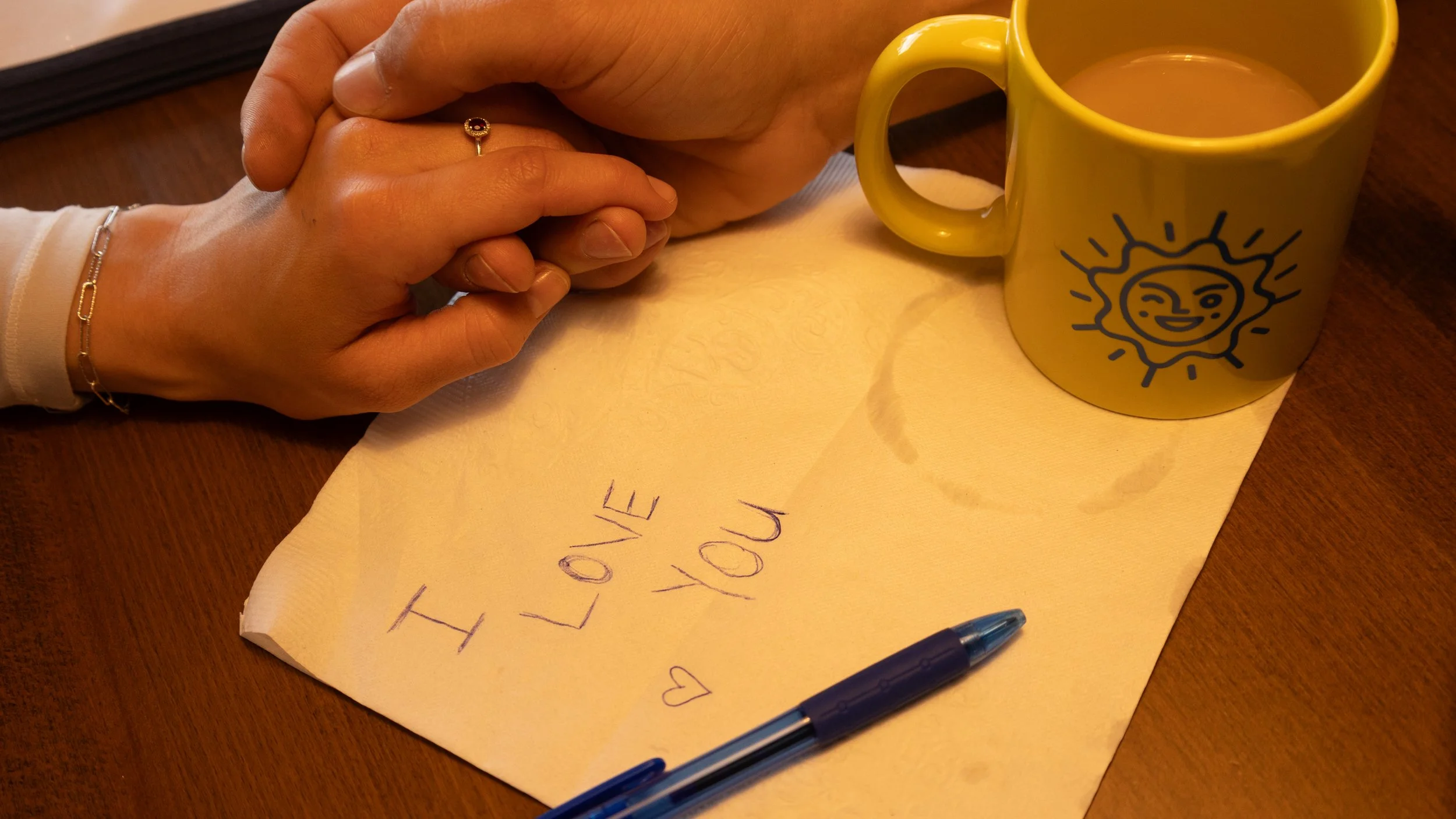 A person is holding another person's hand while sitting at a table. On the table, there is a yellow mug with a sun face drawing, a handwritten note that says 'I Love You' with a heart, and a blue pen.