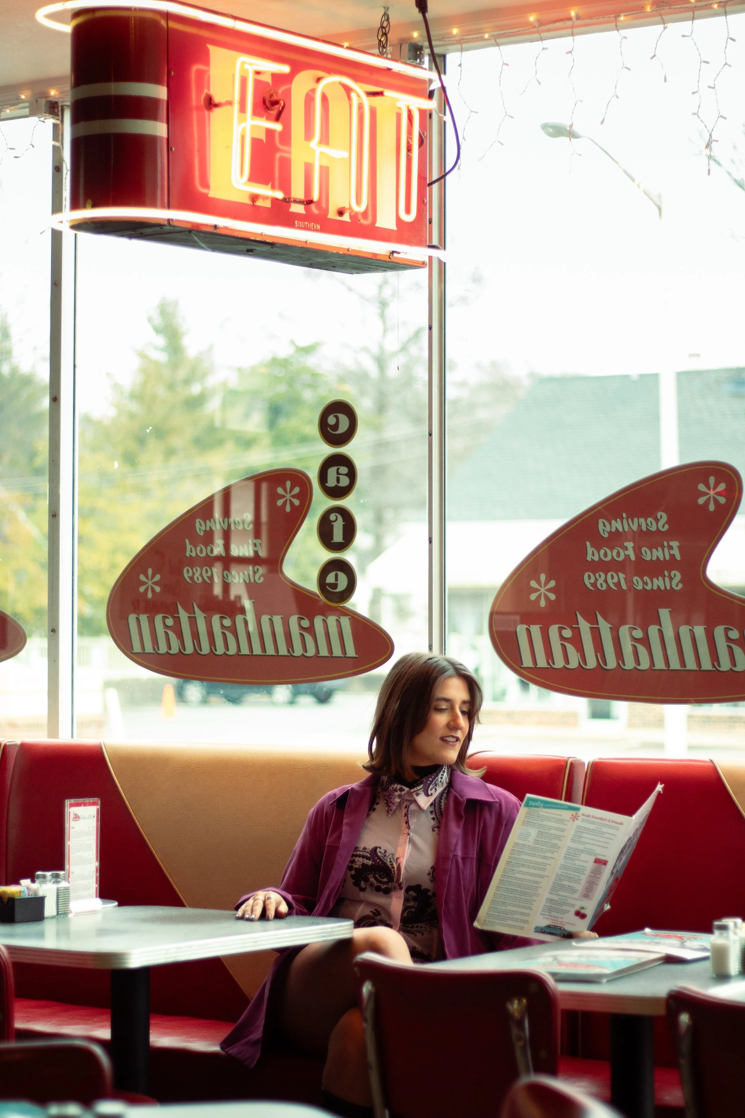 A woman sitting inside a retro diner reads a menu, with red booths, a large window, and illuminated signs that say 'Eat' and 'Manhattan' in the background.