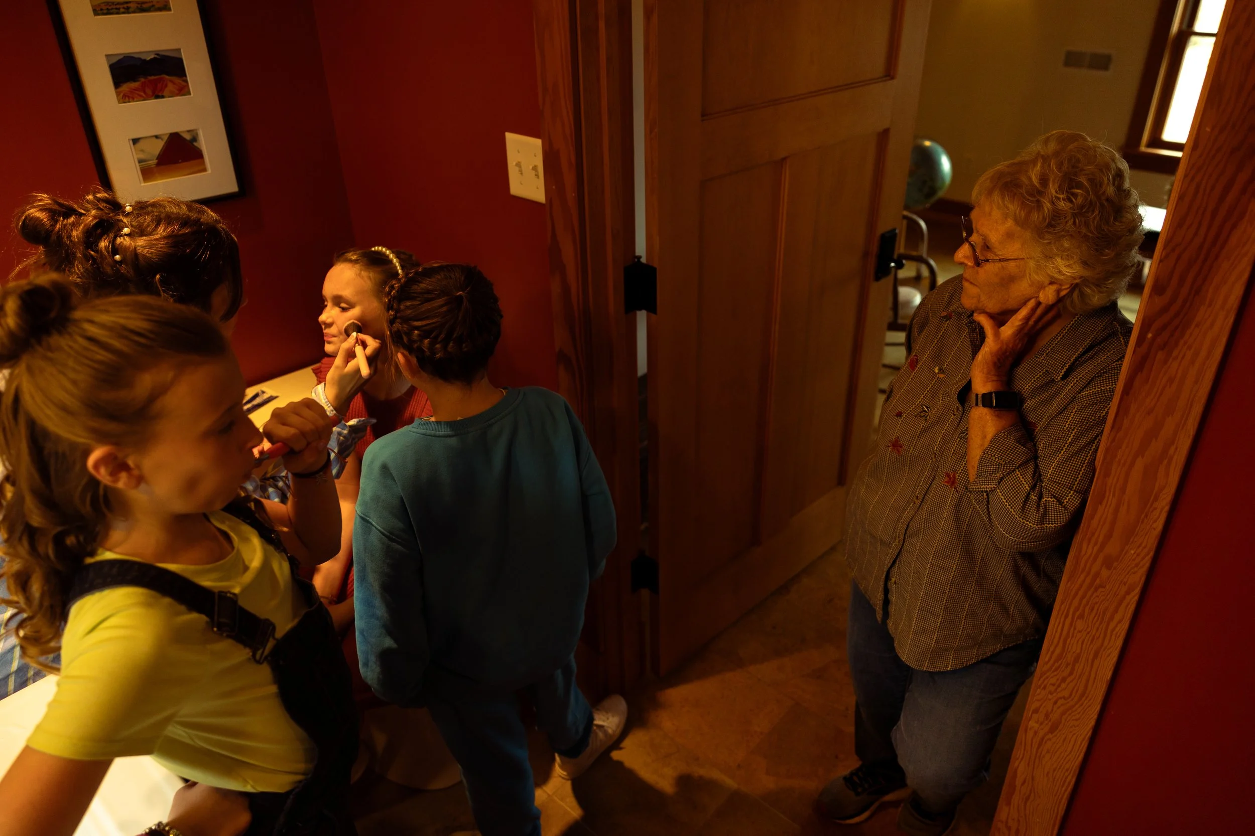A group of young girls getting their faces painted by a woman inside a home or studio, with an older woman watching nearby.