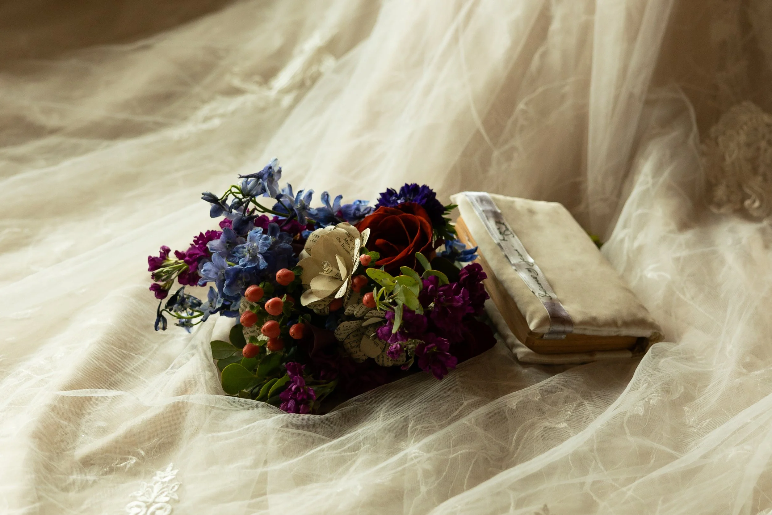 A bouquet of multicolored flowers and a vintage book resting on sheer, cream-colored fabric.
