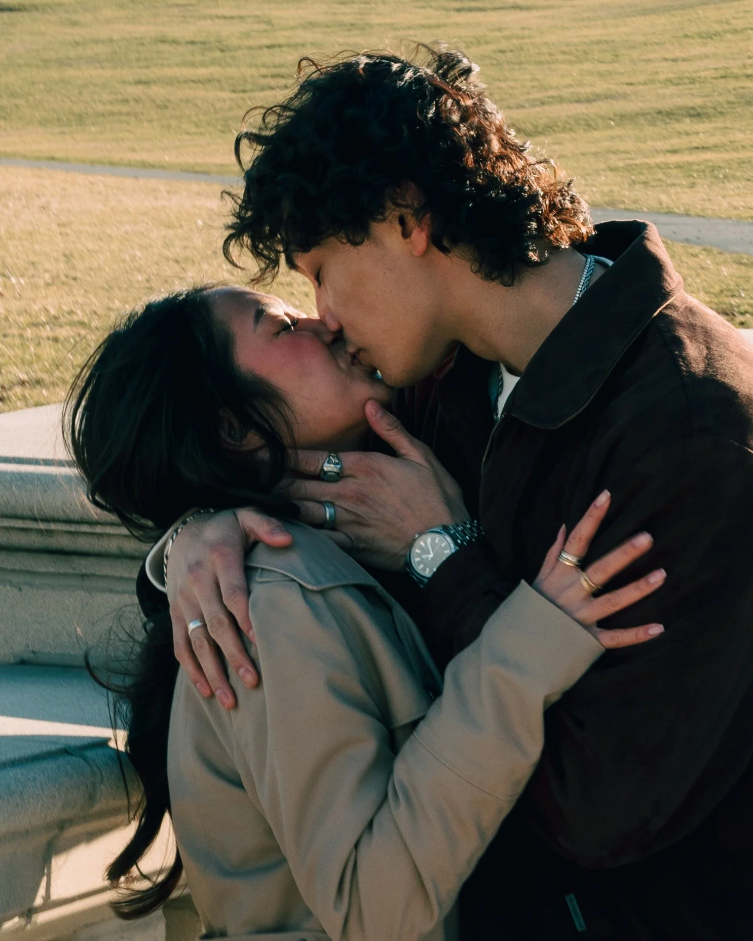A couple passionately kissing outdoors near a park bench with a grassy field in the background.