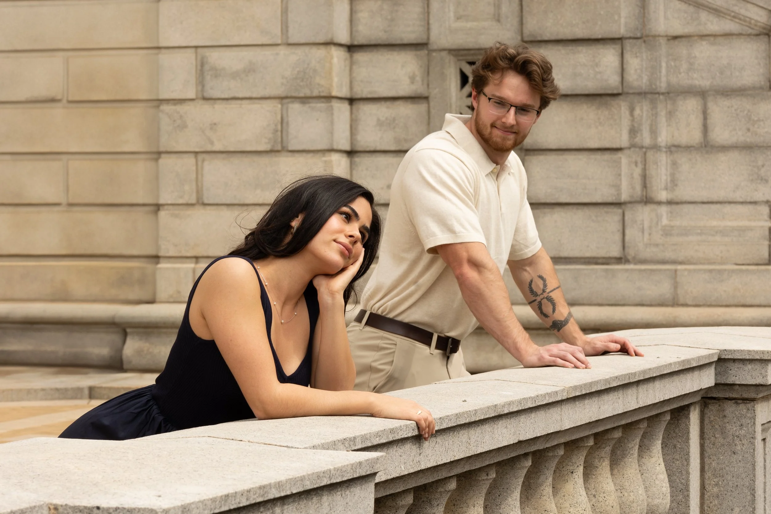 A woman leaning on a stone balcony railing, looking thoughtful or bored, while a man stands beside her, leaning on the railing, looking at her.
