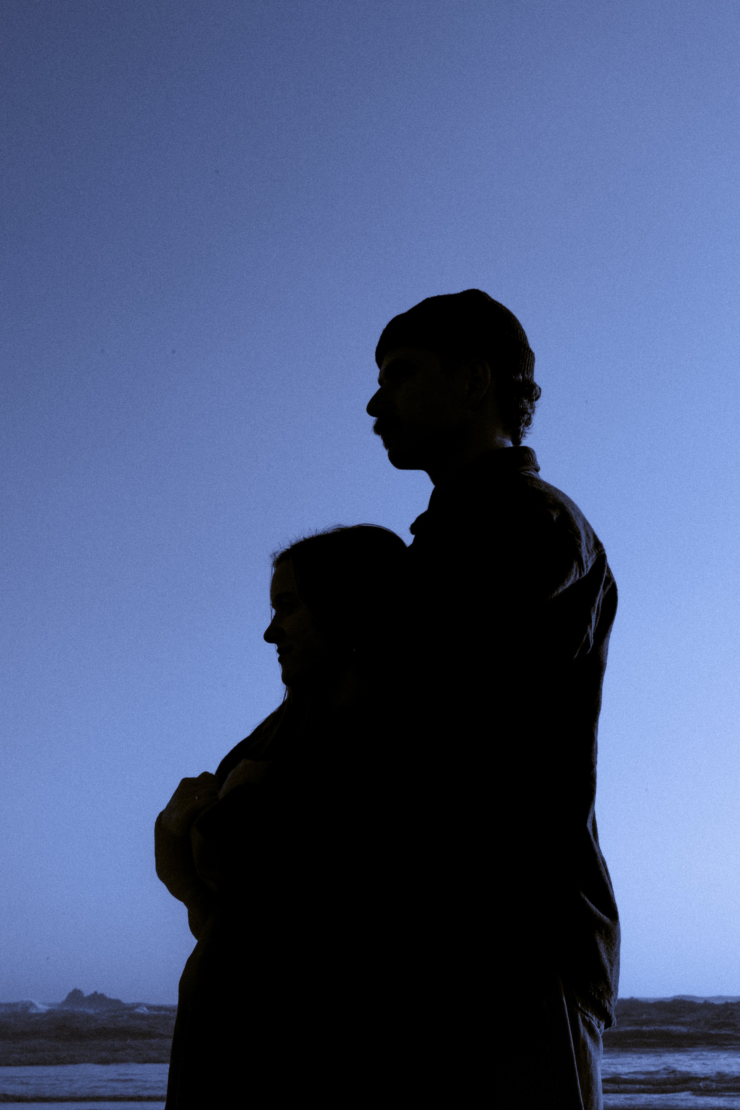 Silhouettes of a man and woman standing close together on a beach at dusk with waves in the background.