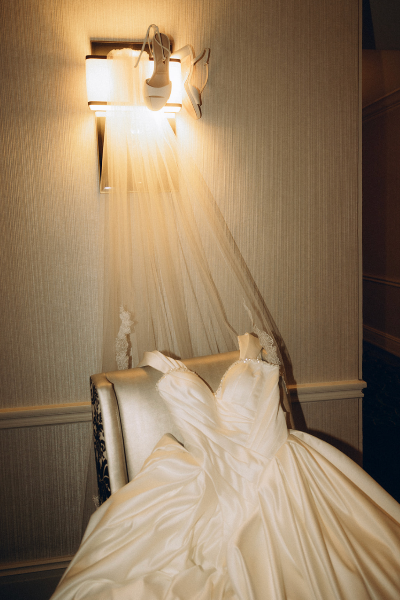 A wedding dress hangs on a mannequin on a chair with a beige wall and a sconce light above.