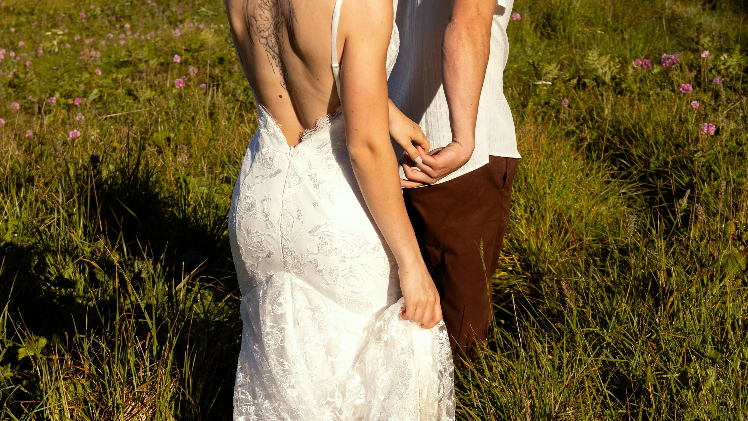 Close-up of a couple holding hands in a grassy field with pink flowers. The woman is wearing a white lace wedding dress, and the man is in a white shirt and brown pants.