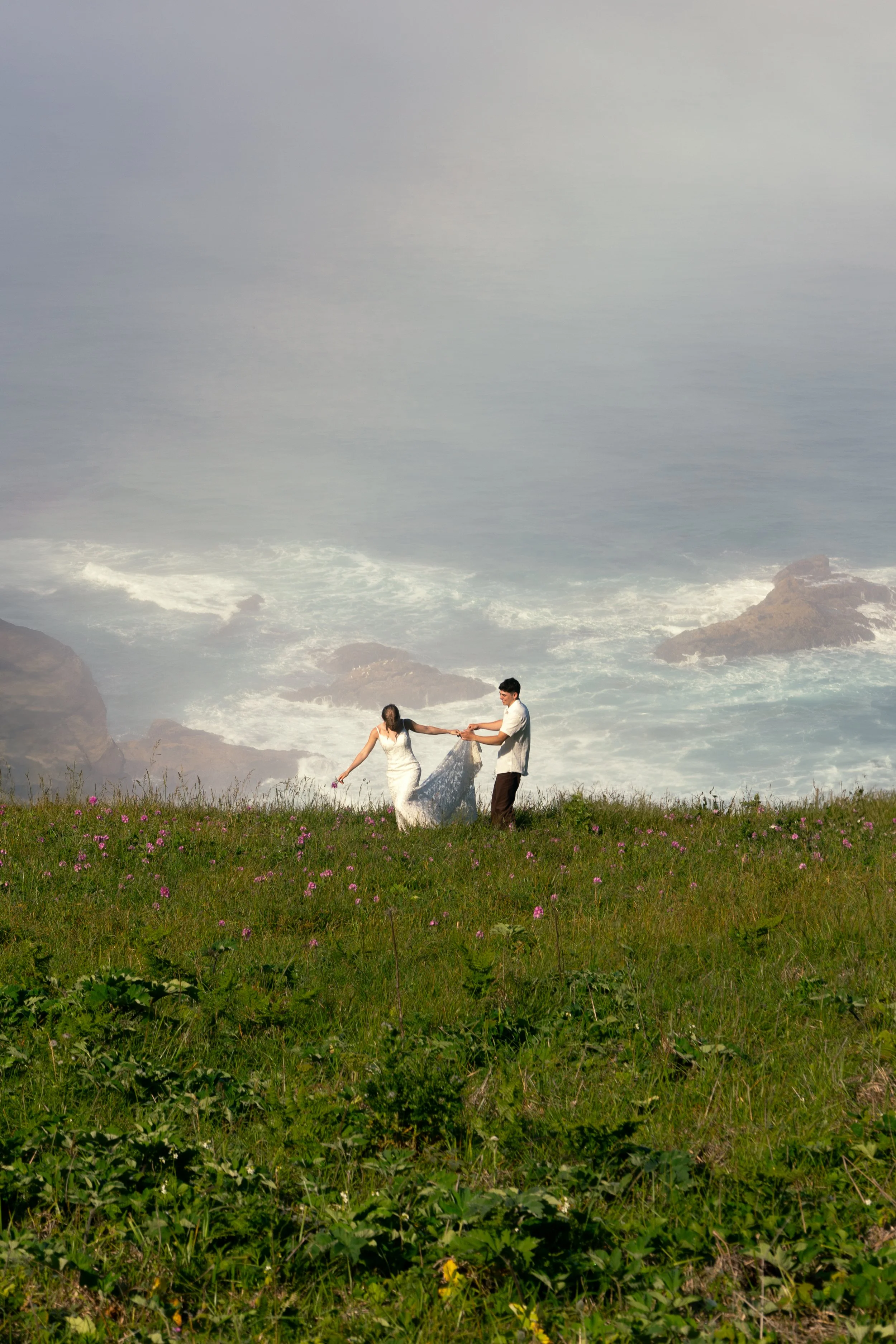 A couple holding hands and playing together on a grassy field with pink flowers, overlooking the ocean with rocks and waves