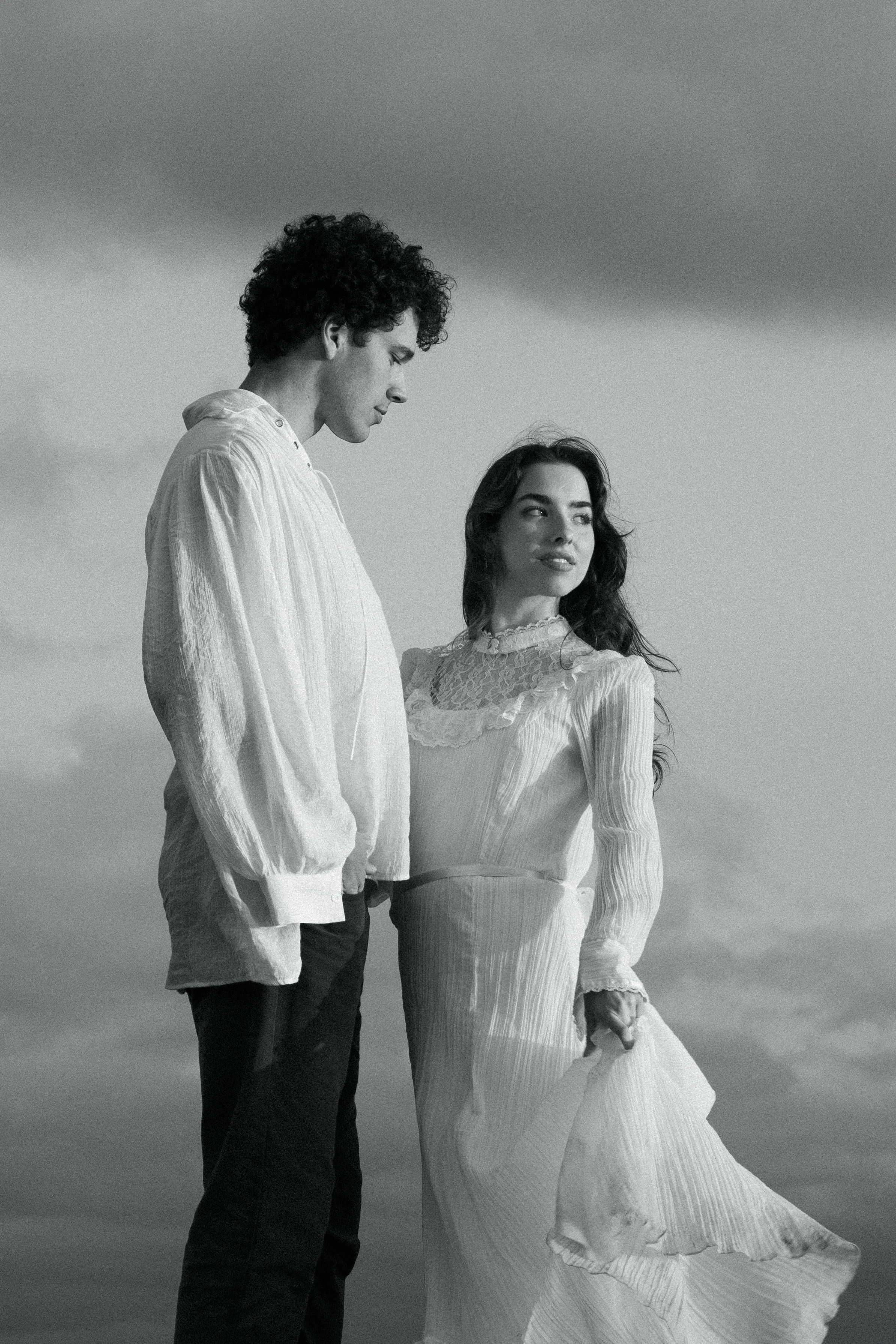 A black and white photo of a young man with curly hair and a woman with long hair, both dressed in vintage-style clothing, standing outdoors under a cloudy sky.
