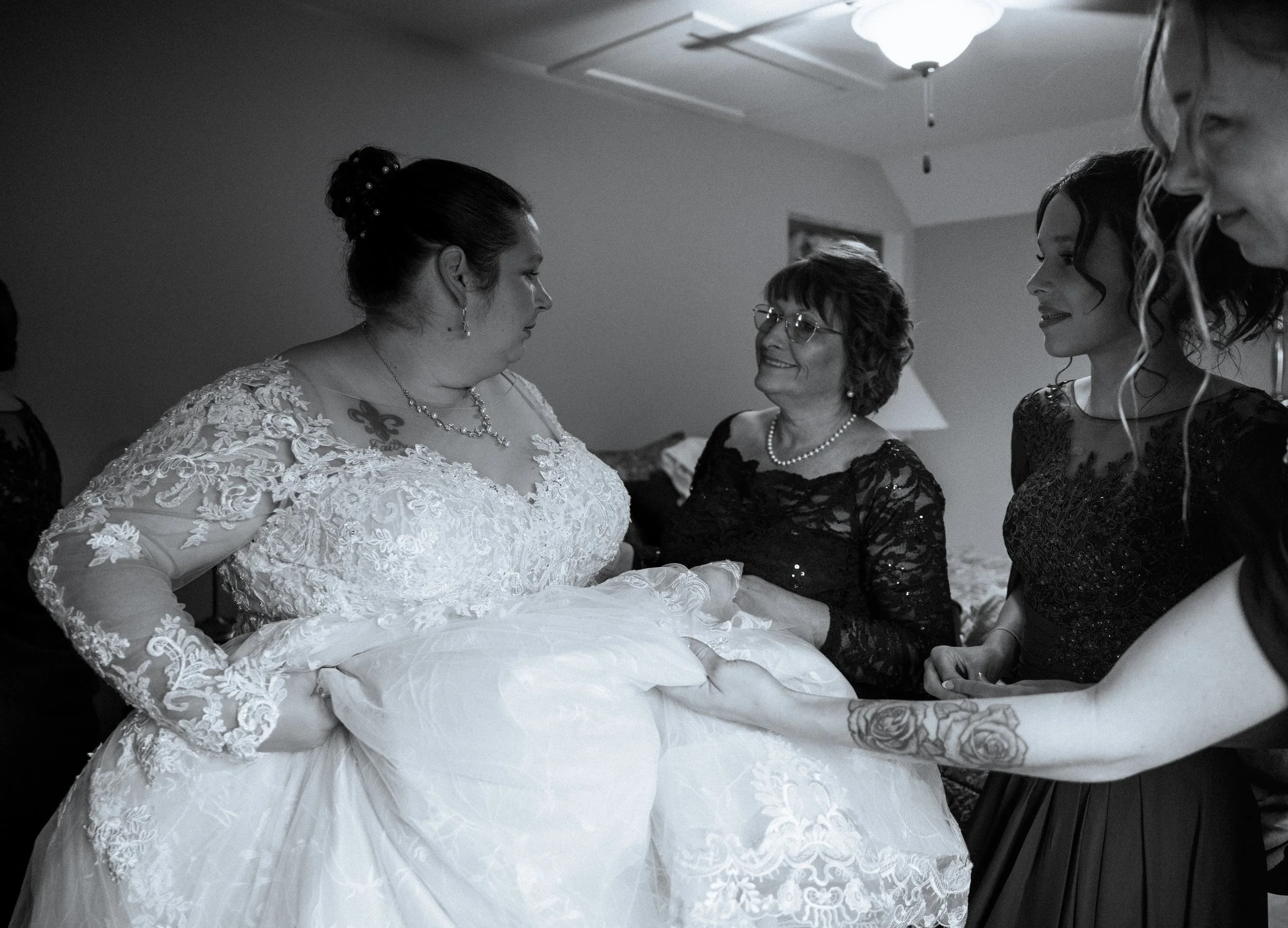 A bride in a wedding dress receiving assistance from women in black lace dresses in a room with a ceiling light.