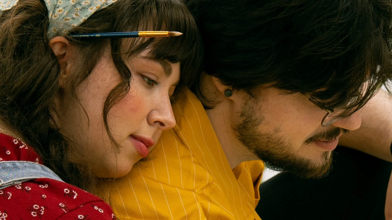 A woman with dark curly hair wearing a red top with flower prints and a denim overall, resting her head on the shoulder of a man with dark hair, glasses, and a beard, wearing a yellow shirt.