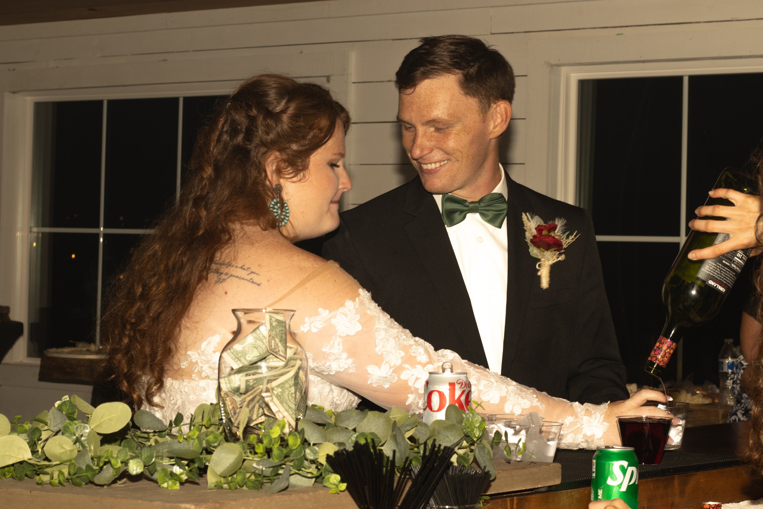 A bride and groom smiling at each other during a wedding reception. They are dressed in formal attire, with the bride wearing a lace wedding dress and the groom in a tuxedo with a green bow tie and boutonniere.