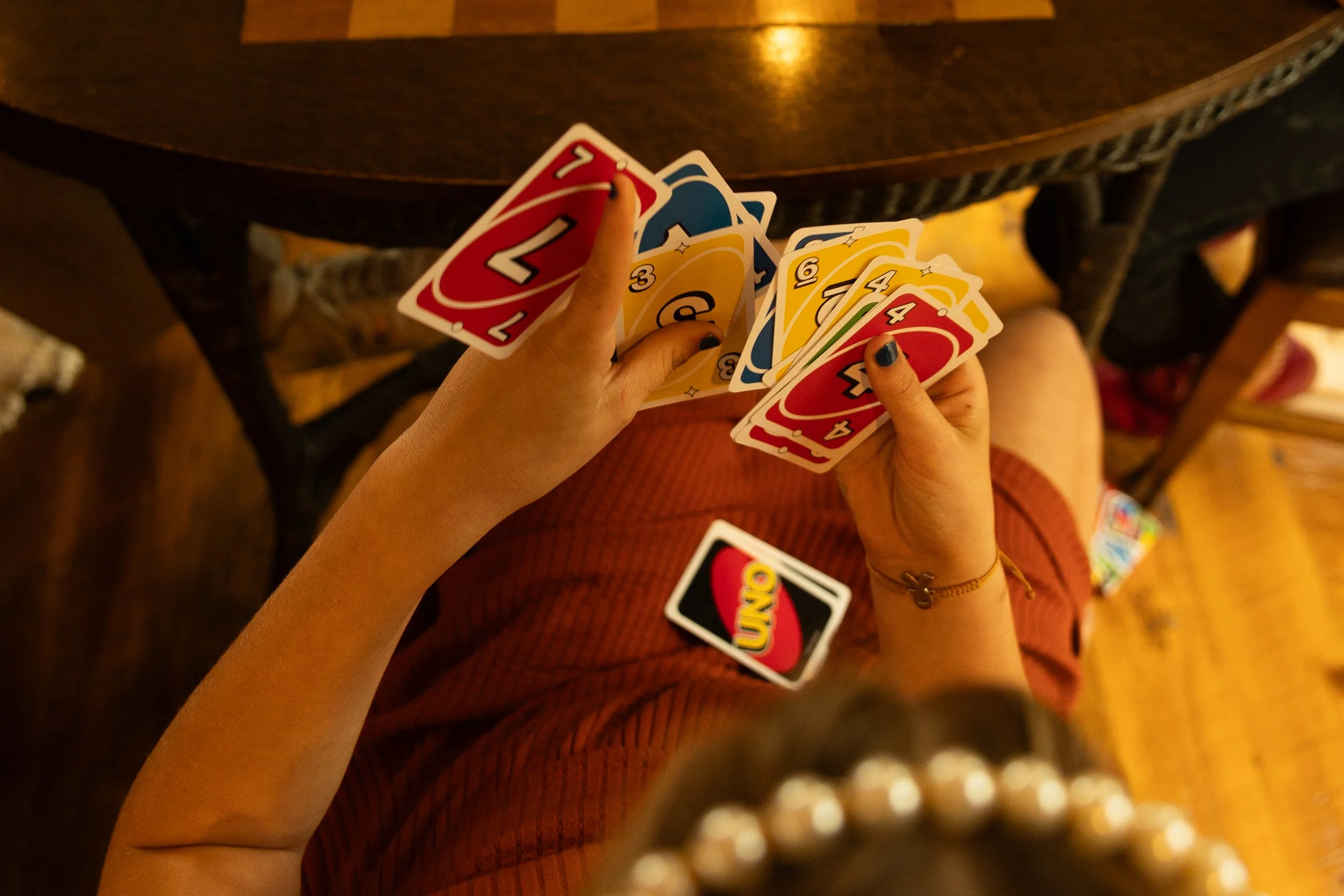 Person holding UNO cards, sitting at a wooden table, with other UNO cards spread on their lap and on the table, in a cozy indoor setting.