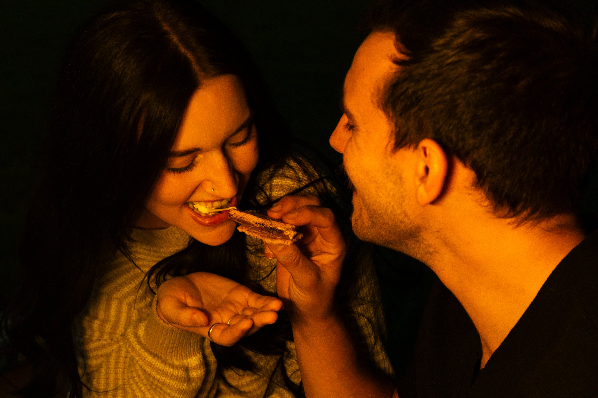 A woman with long dark hair wearing a beige sweater is about to eat a piece of chocolate cake given by a man with short dark hair, wearing a black shirt, both smiling and in a warm, intimate setting.