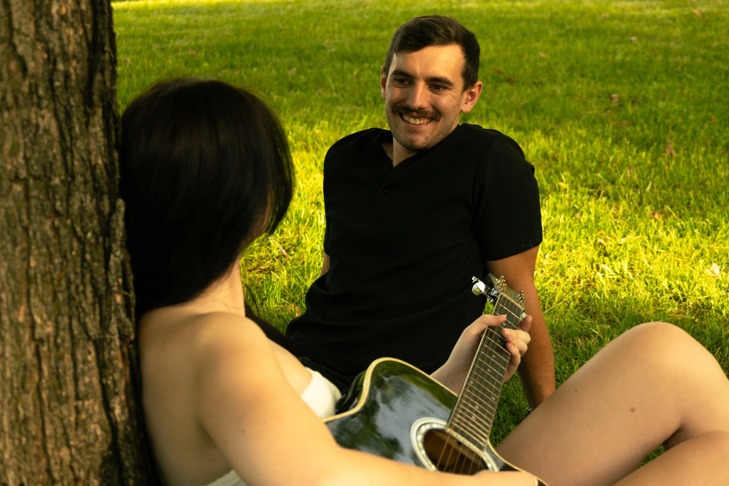 A woman with dark hair sitting on the grass and leaning against a tree, holding an acoustic guitar, while a man with short dark hair and a beard, wearing a black shirt, sits nearby on the grass smiling.