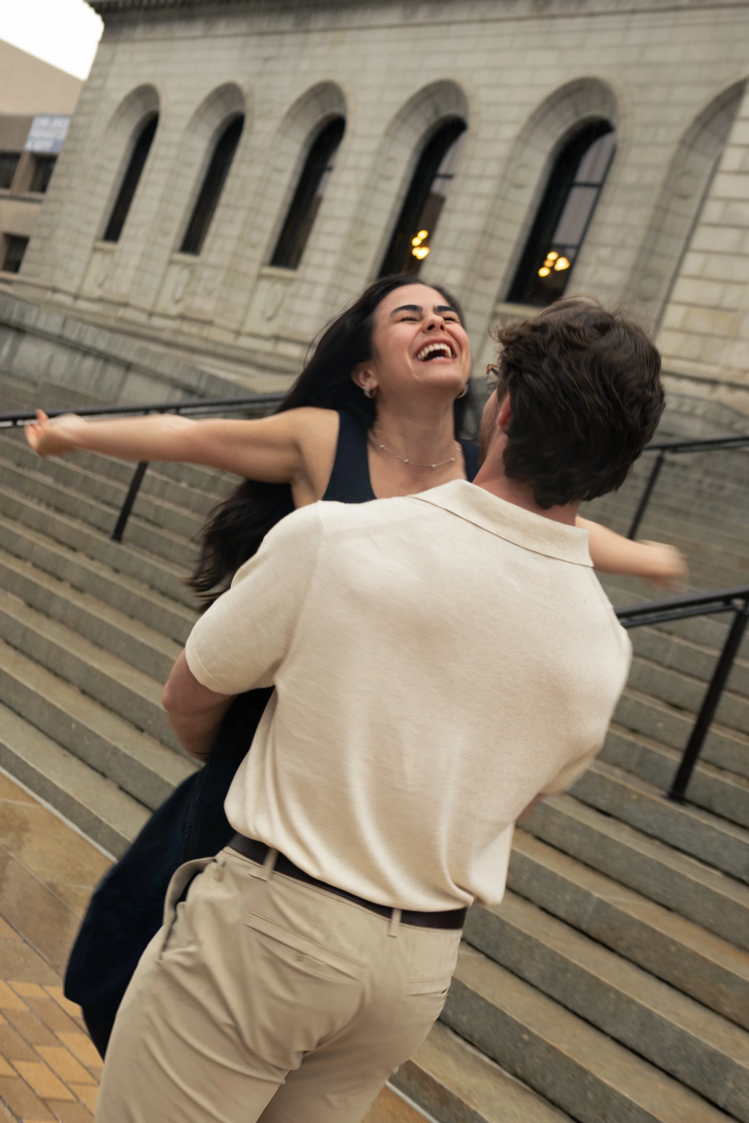 A woman with long dark hair joyful being lifted by a man in front of a building with arched windows and stairs.