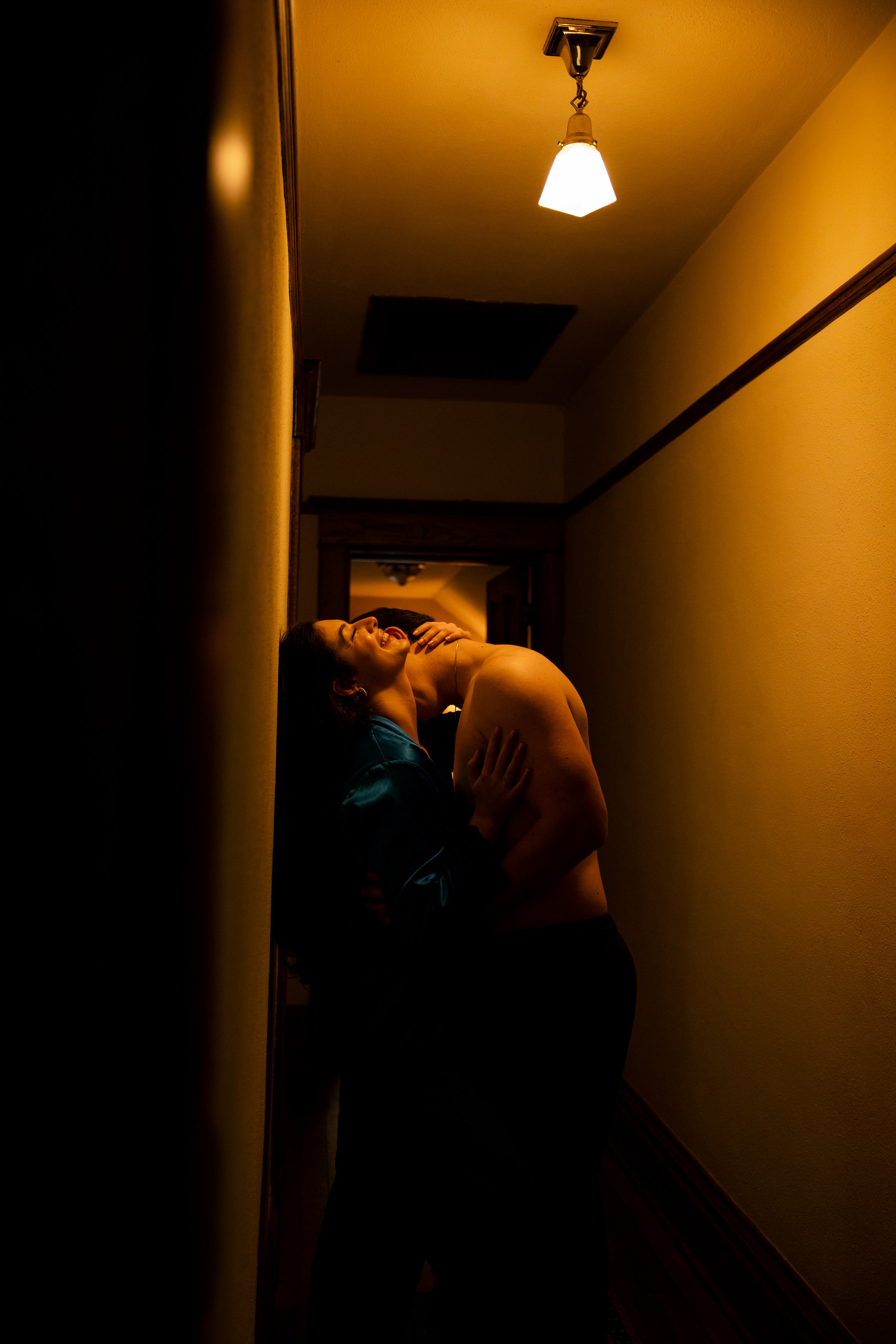 A woman leaning against the wall, laughing with her eyes closed, as a man wraps his arms around her in a hallway illuminated by a ceiling light.