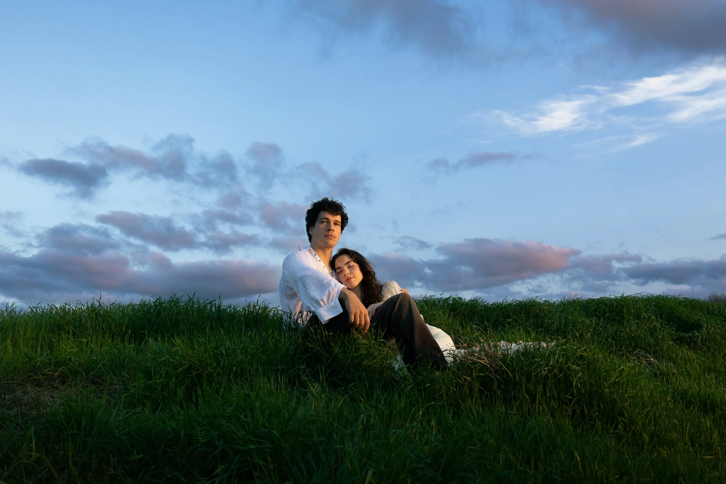 A young man and woman sitting on green grass with a blue sky and clouds in the background. The woman is resting her head on the man's shoulder, and they both appear calm and relaxed.