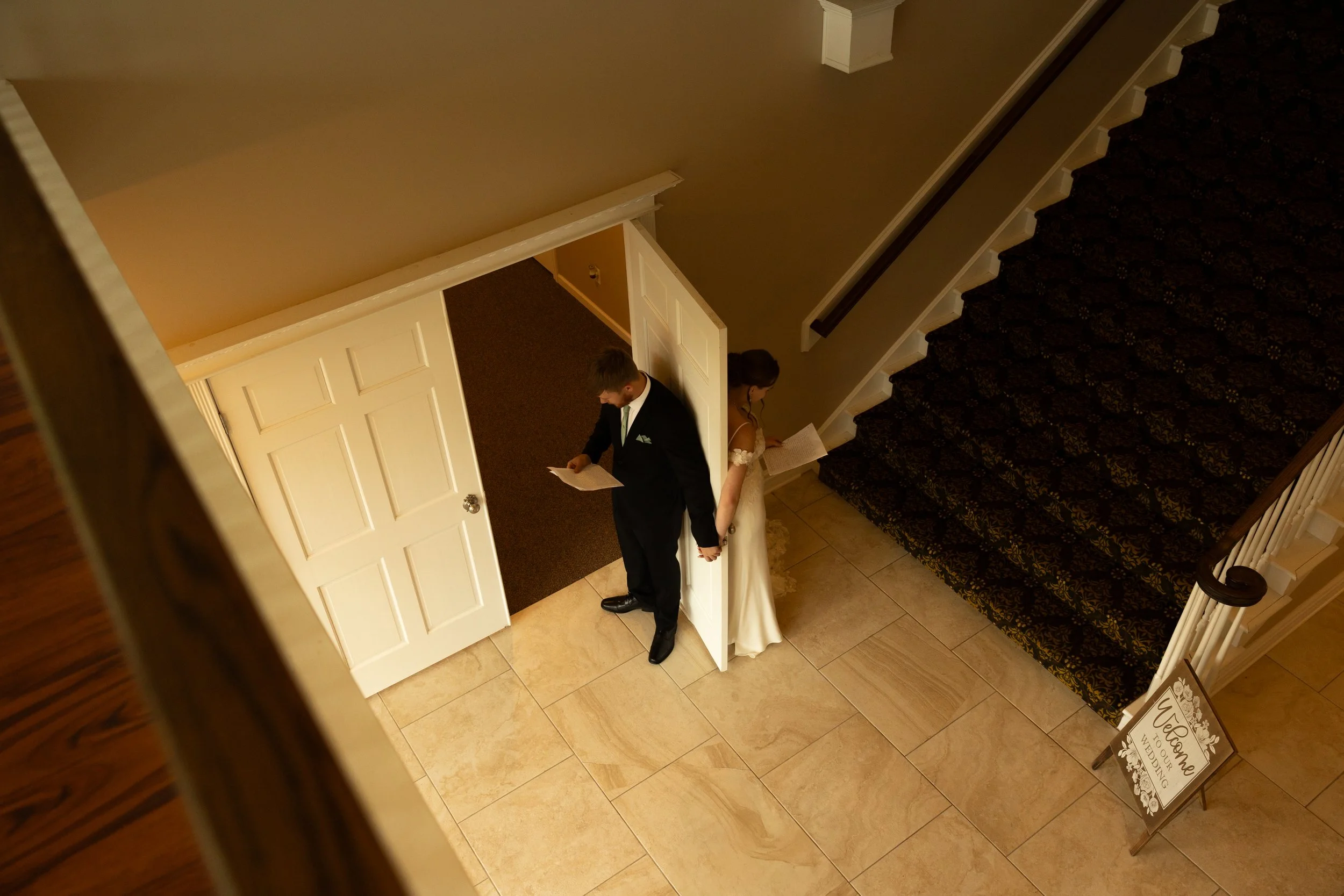 A bride and groom standing back to back, holding hands, and looking at papers in an entrance hall with staircase and a 'Welcome' sign.